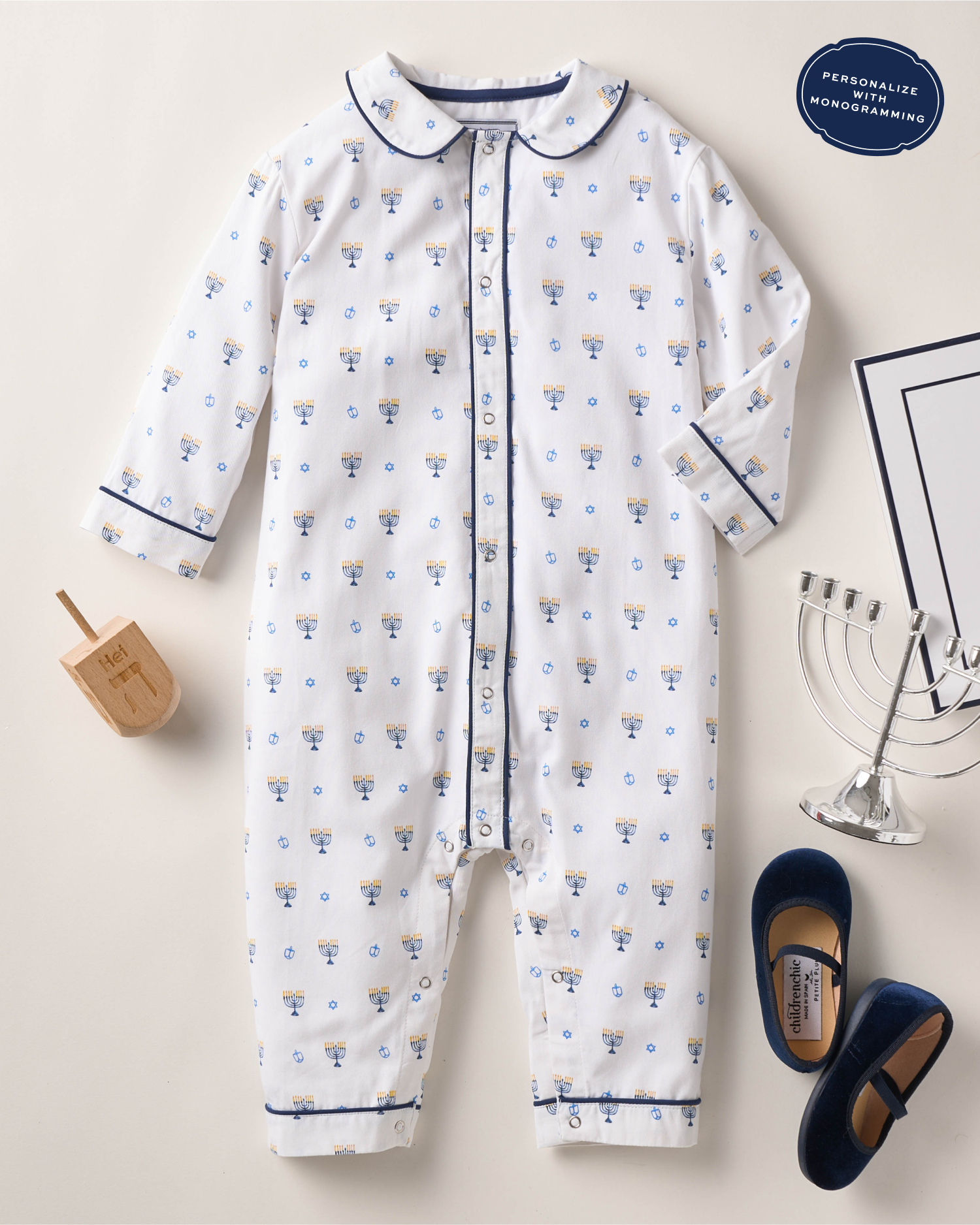 White baby onesie with blue menorah print, surrounded by a wooden dreidel, a silver menorah, blue baby shoes, and a paper. A label reads “Personalize with Monogramming” in the top right corner.