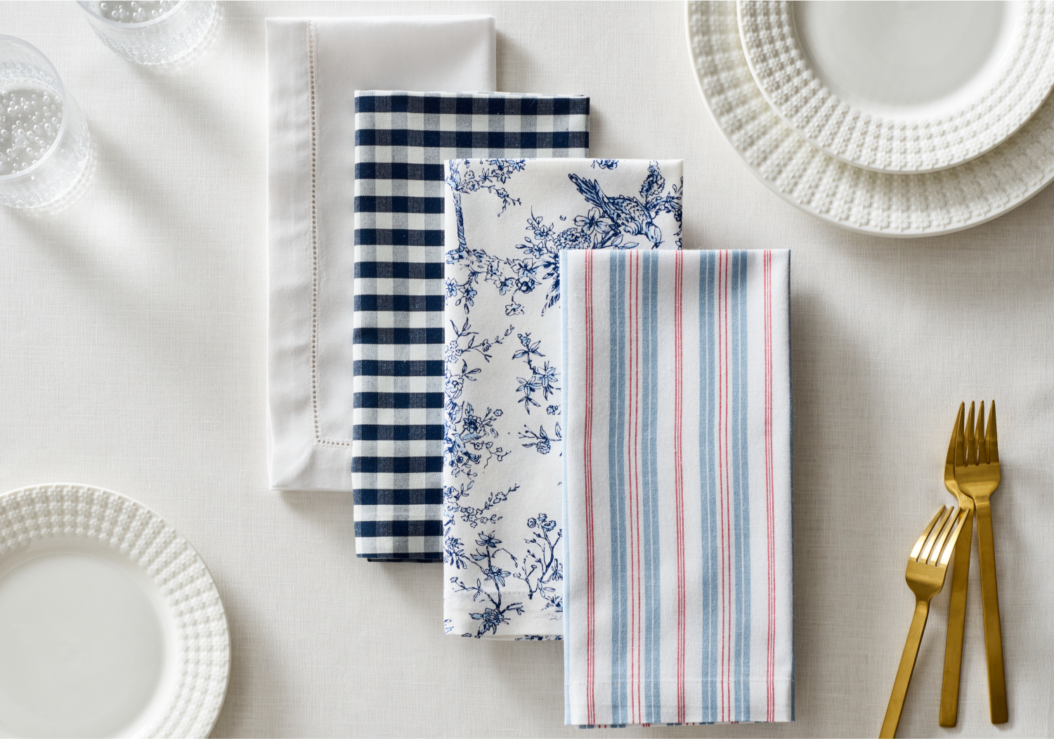 Four neatly folded cloth napkins with different patterns—white with stitching, blue gingham, blue floral, and red-blue striped—arranged on a white tablecloth with plates, glasses, and gold flatware.