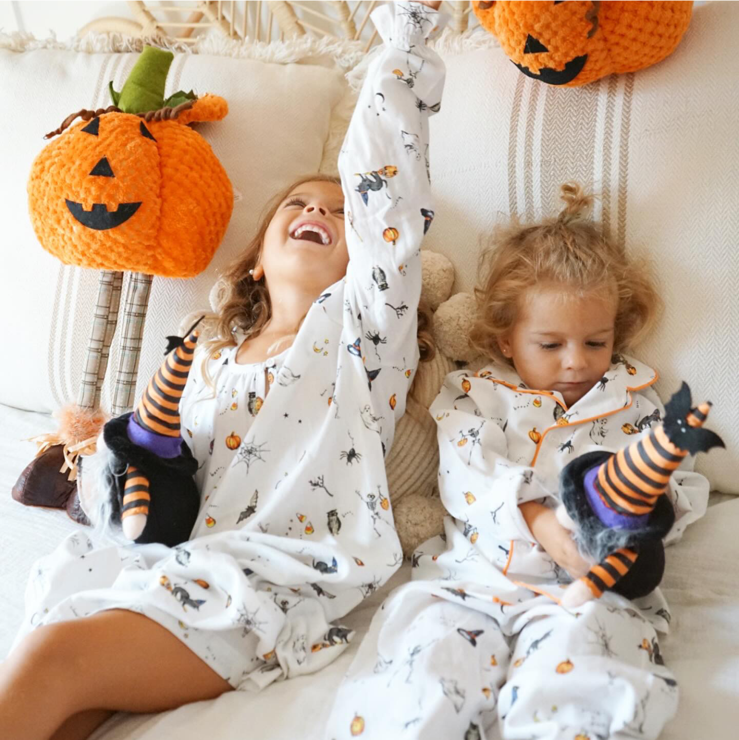 Two young children in Halloween-themed pajamas sit on a couch with plush witch dolls. One child laughs joyfully with an arm raised, while the other looks down. Orange pumpkin decorations are on the couch behind them.
