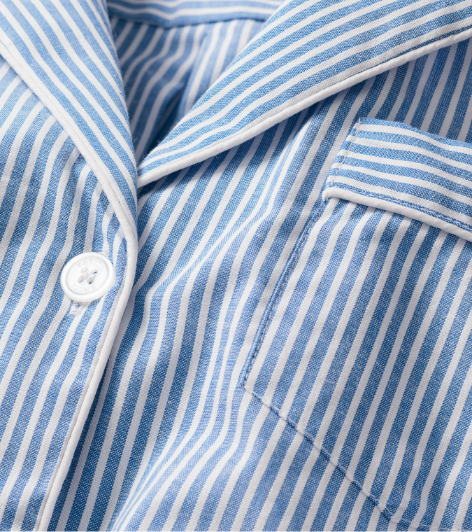Close-up of a blue and white vertically striped shirt with white piping, a white button, and a chest pocket. The fabric appears soft and smooth, resembling classic pajama material.