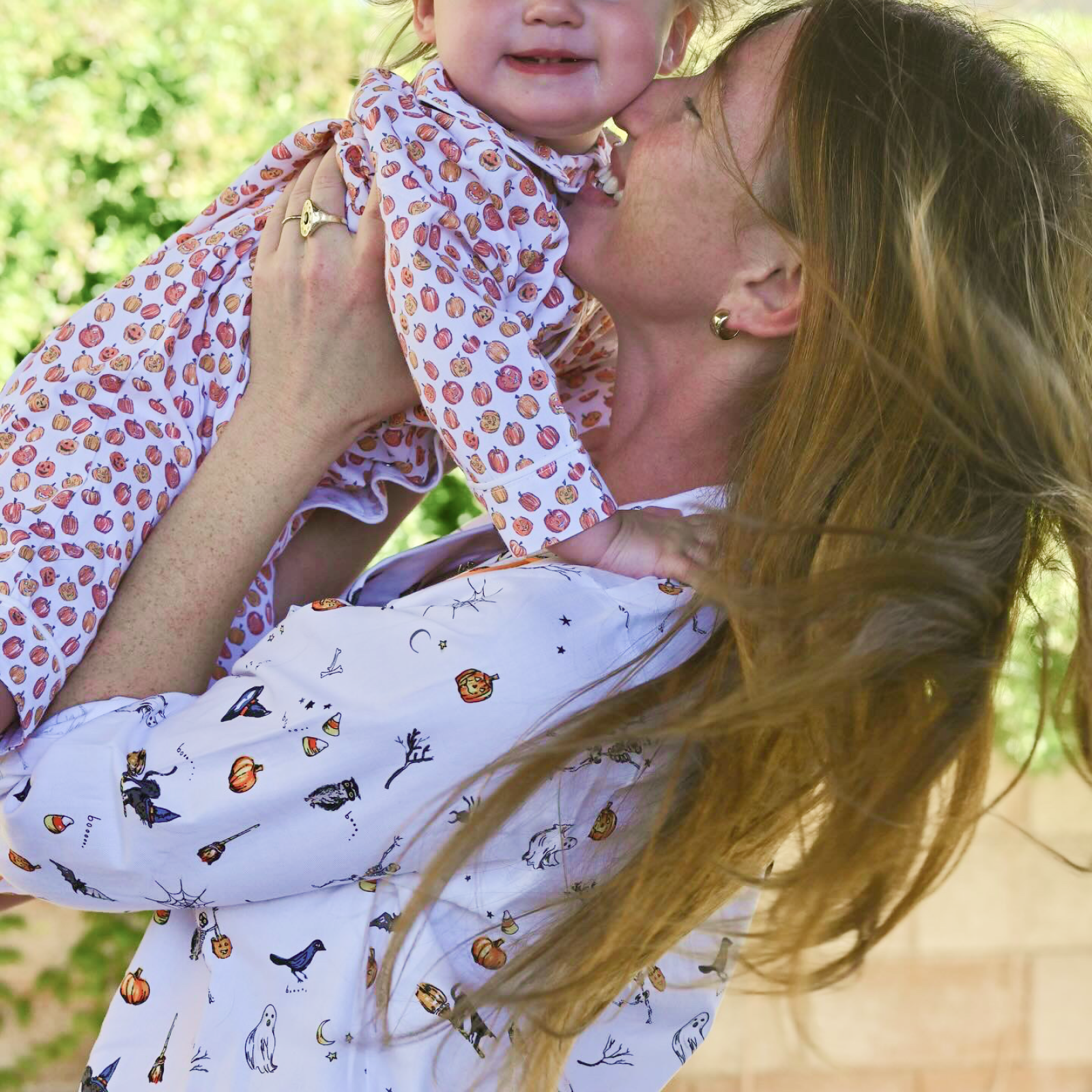 A woman with long hair lifts a smiling baby in the air outdoors. Both wear patterned outfits, and the background is filled with greenery and a brick wall. The woman appears to be kissing the childs cheek.