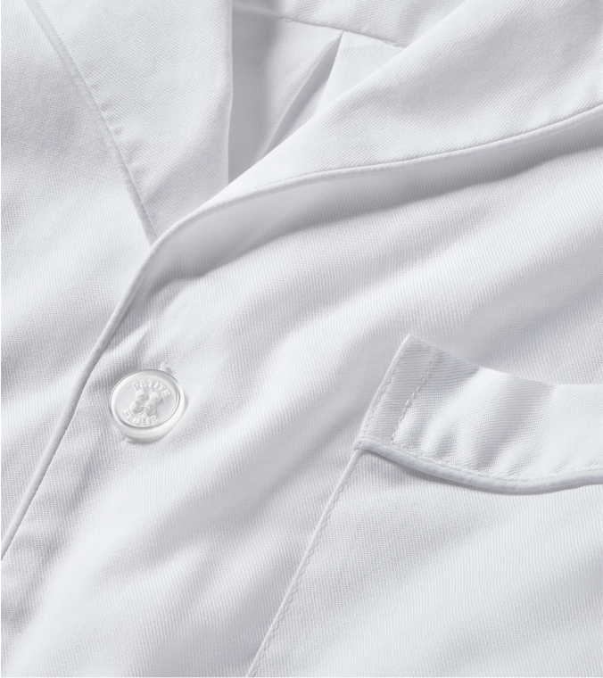 Close-up of a white button-up shirt showing part of the collar, a single button, and a chest pocket. The fabric appears smooth and crisp.