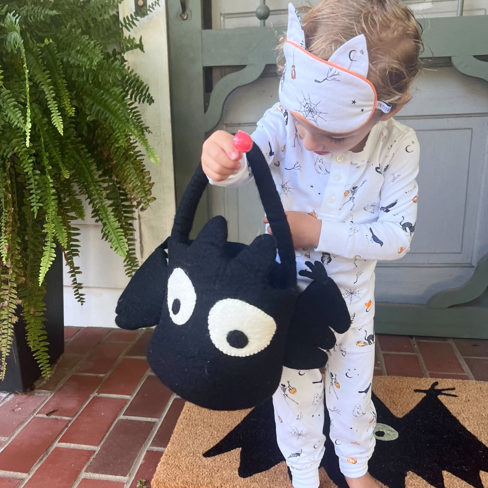 A young child in Halloween pajamas and a cat-ear headband stands on a porch, holding a black bat-shaped trick-or-treat basket and looking inside. A green fern and doormat are nearby.