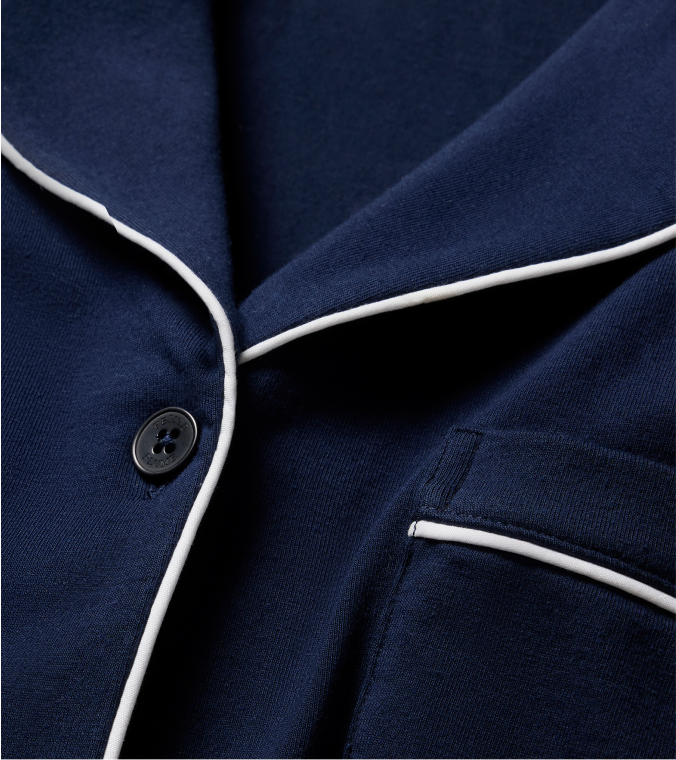 Close-up of a navy blue pajama top with white piping along the collar and pocket edge, and a single black button visible. The fabric appears soft and smooth.