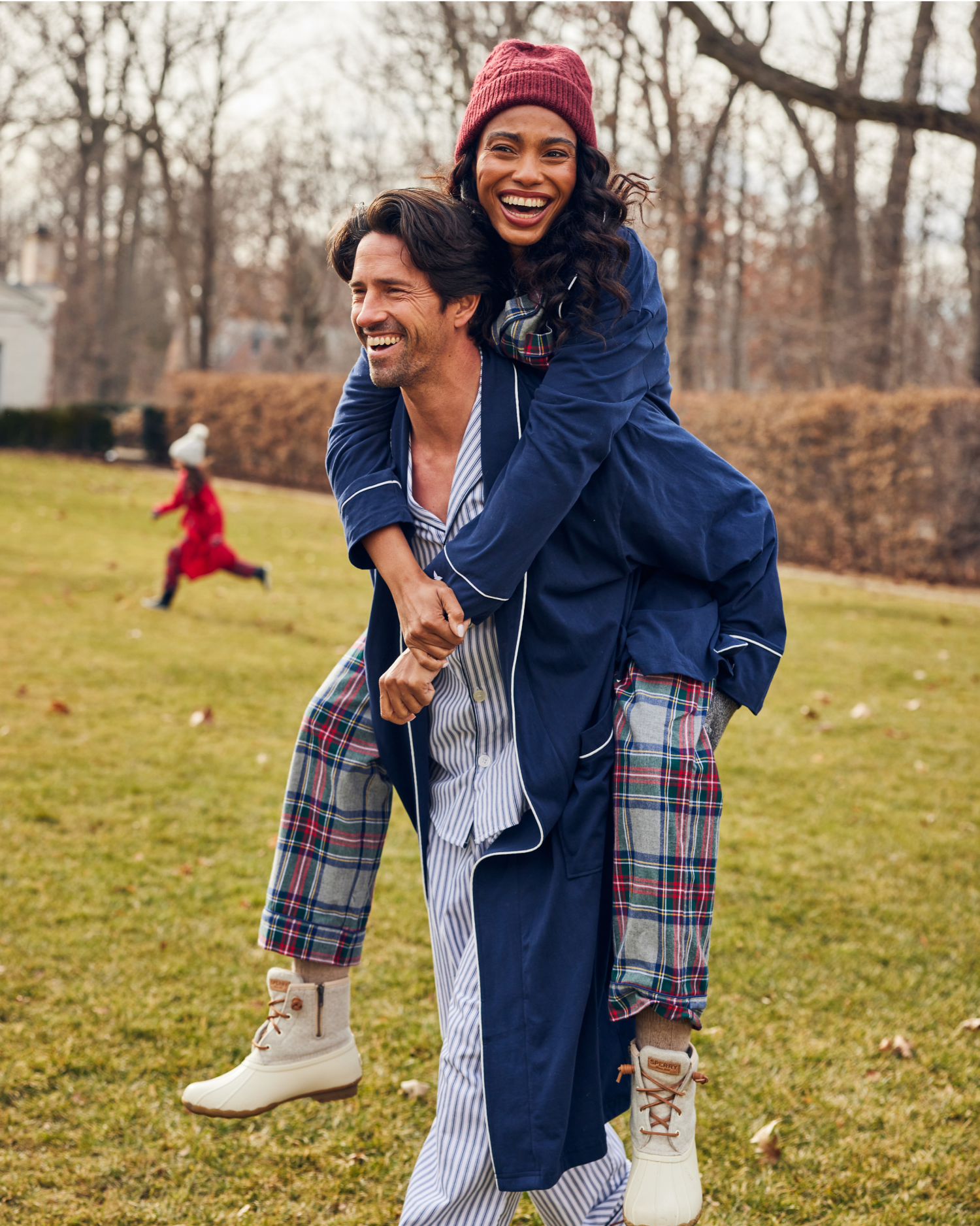 A smiling woman in pajamas and a red beanie rides piggyback on a man, also in pajamas, outside on a grassy lawn. Another person in red runs in the background among leafless trees.