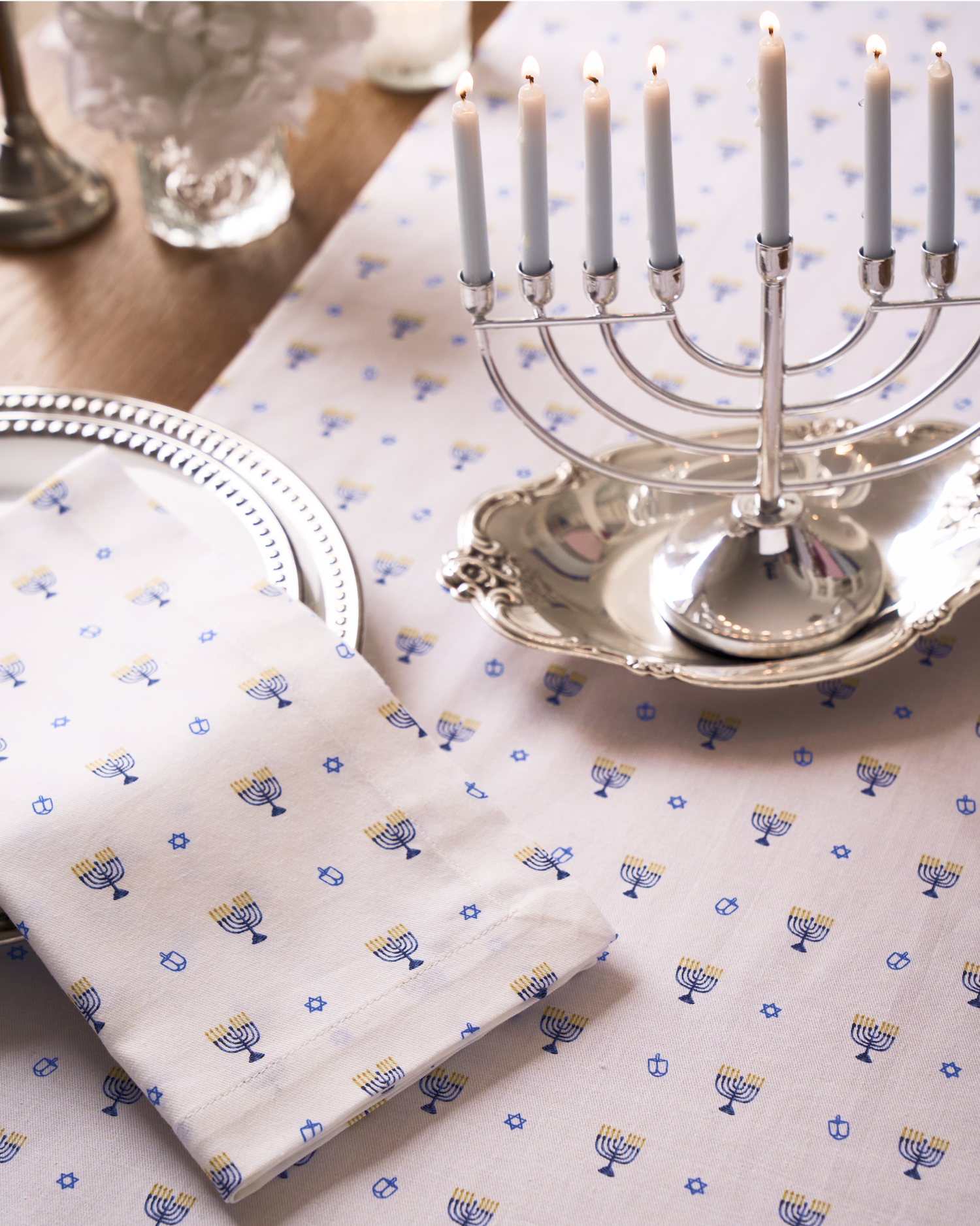 A silver menorah with lit candles sits on a silver tray atop a white tablecloth decorated with small blue and yellow menorah patterns. A matching napkin and a silver plate are set nearby.