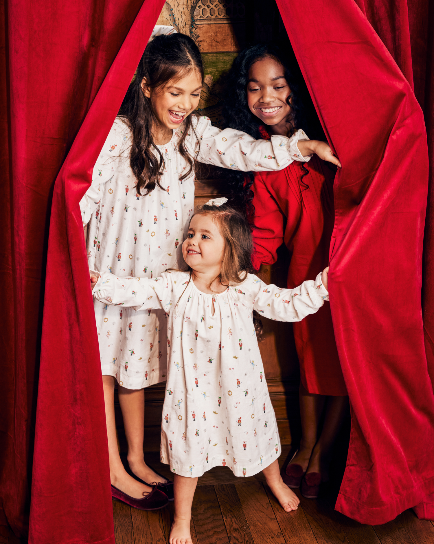 Three young girls smile and laugh as they stand between red velvet curtains. Two girls in matching white dresses with prints hold the curtains open, while a third girl in a red dress stands behind them on a wooden floor.