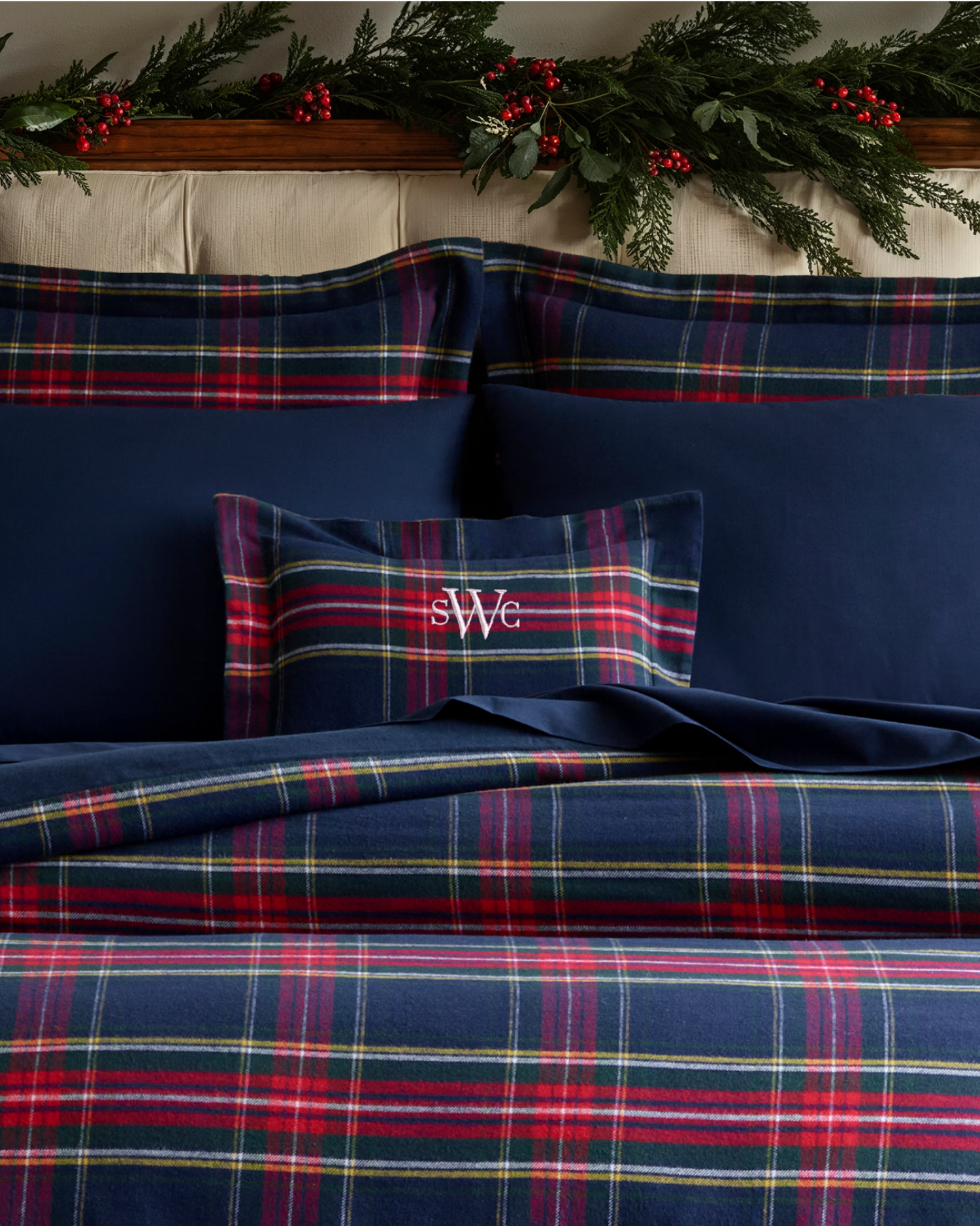 A bed with navy blue and red plaid bedding, a navy blue pillow with white monogram sWe, and festive greenery with red berries decorating the headboard.