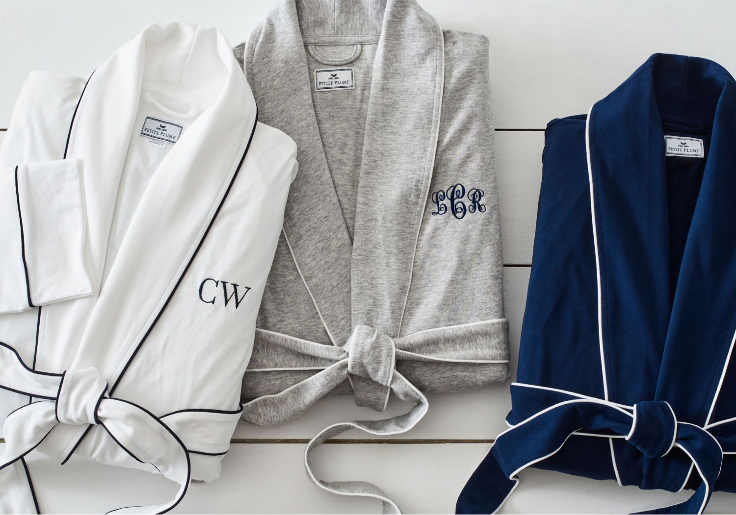 Three bathrobes are neatly folded side by side: one white with black trim and “CW” initials, one light gray with blue “ECB” initials, and one solid navy blue, all tied at the waist with matching belts.