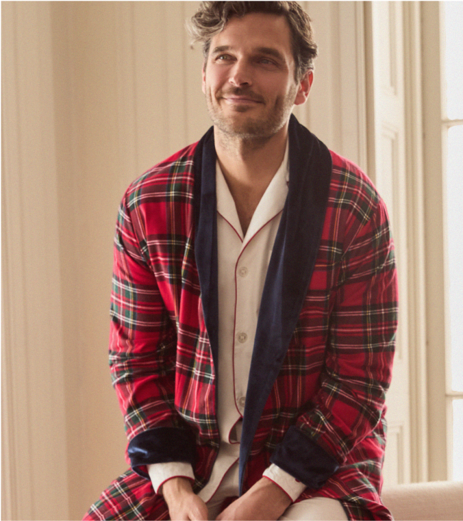 A man with wavy hair smiles softly while sitting indoors, wearing a red plaid robe with dark lapels over light-colored pajamas. Warm natural light streams through a window behind him.