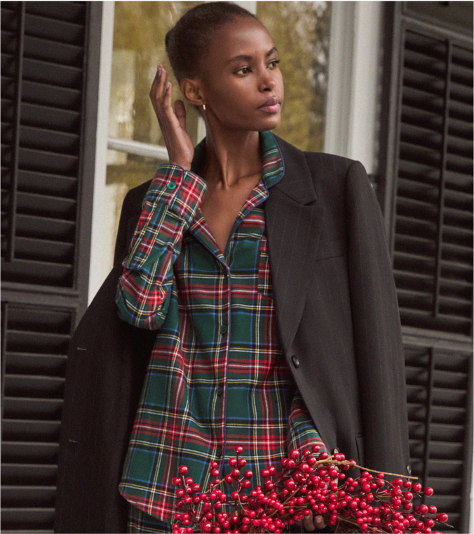 A woman wearing a black blazer draped over her shoulders and a green plaid shirt stands in front of dark shutters, holding a bunch of red berries and looking to the side with one hand near her ear.