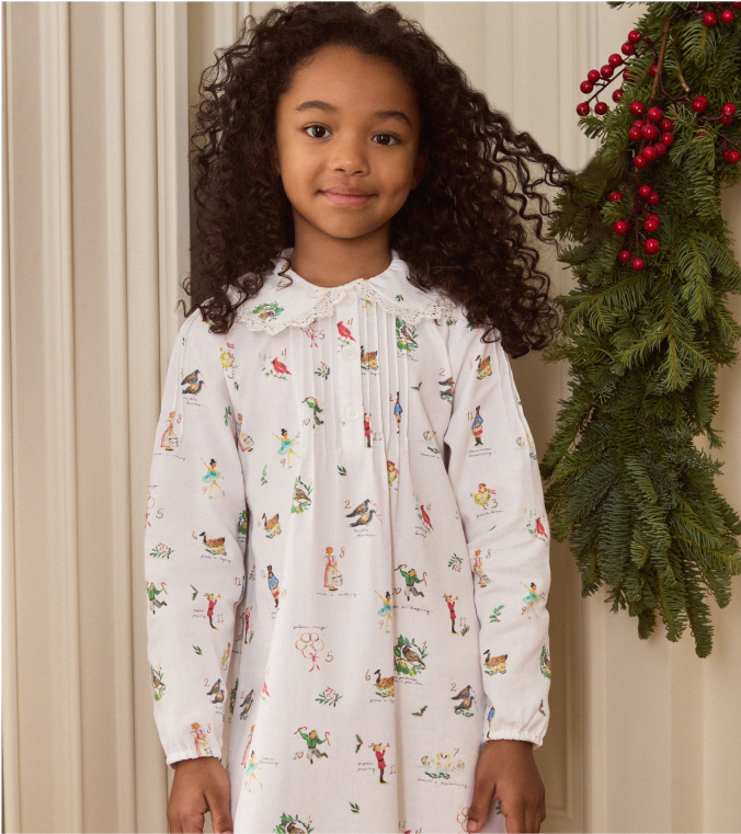 Young girl with long curly hair wearing a white dress with colorful holiday prints, standing indoors next to green pine branches and red berries.