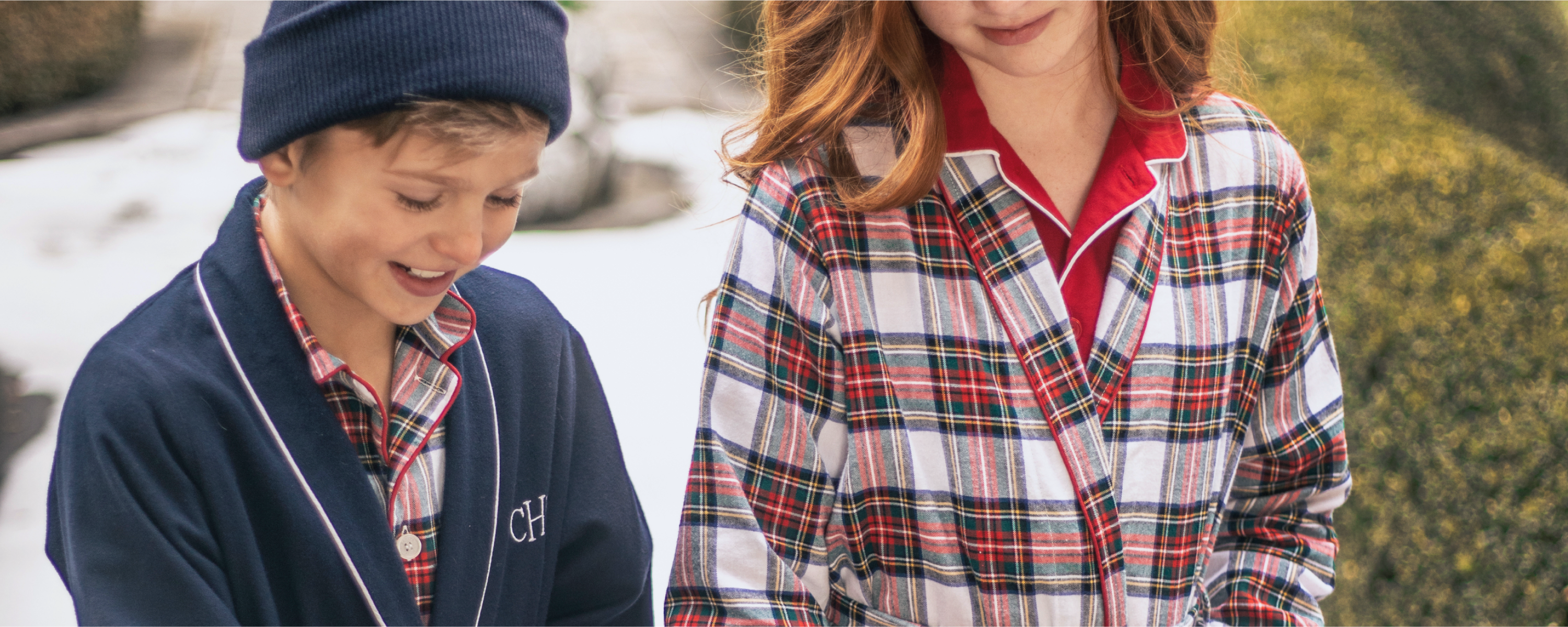 A boy in a blue beanie and navy robe with initials CH smiles beside a girl with red hair wearing a plaid robe and red shirt; both stand outdoors with greenery and snow in the background. A boy in a blue beanie and navy robe with initials CH smiles beside a girl with red hair wearing a plaid robe and red shirt; both stand outdoors with greenery and snow in the background.