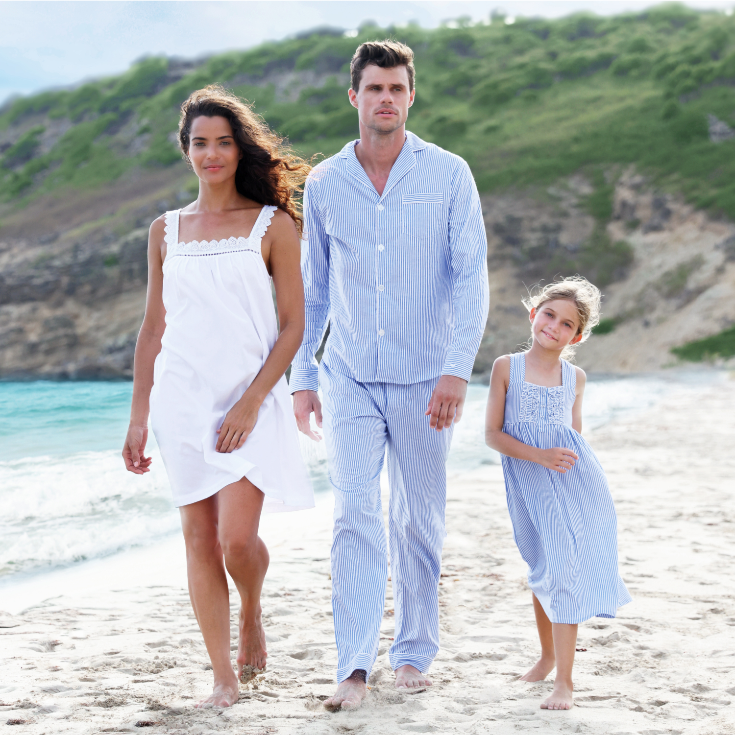 A woman, man, and young girl wearing light-colored sleepwear walk barefoot together along a sandy beach with green hills and ocean waves in the background.