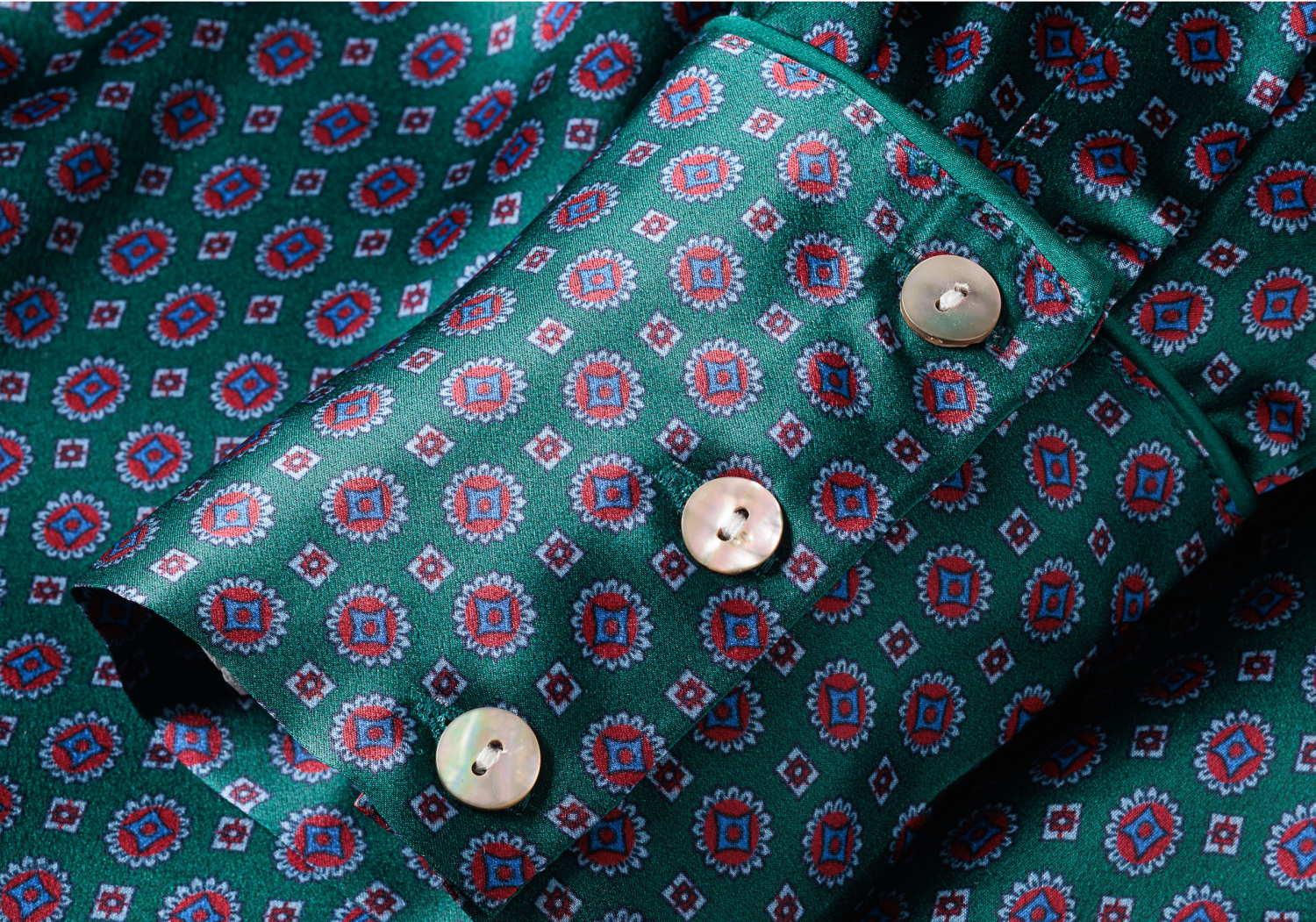 A close-up of a green shirt cuff with a red and blue geometric pattern, featuring three white buttons and stitching details.
