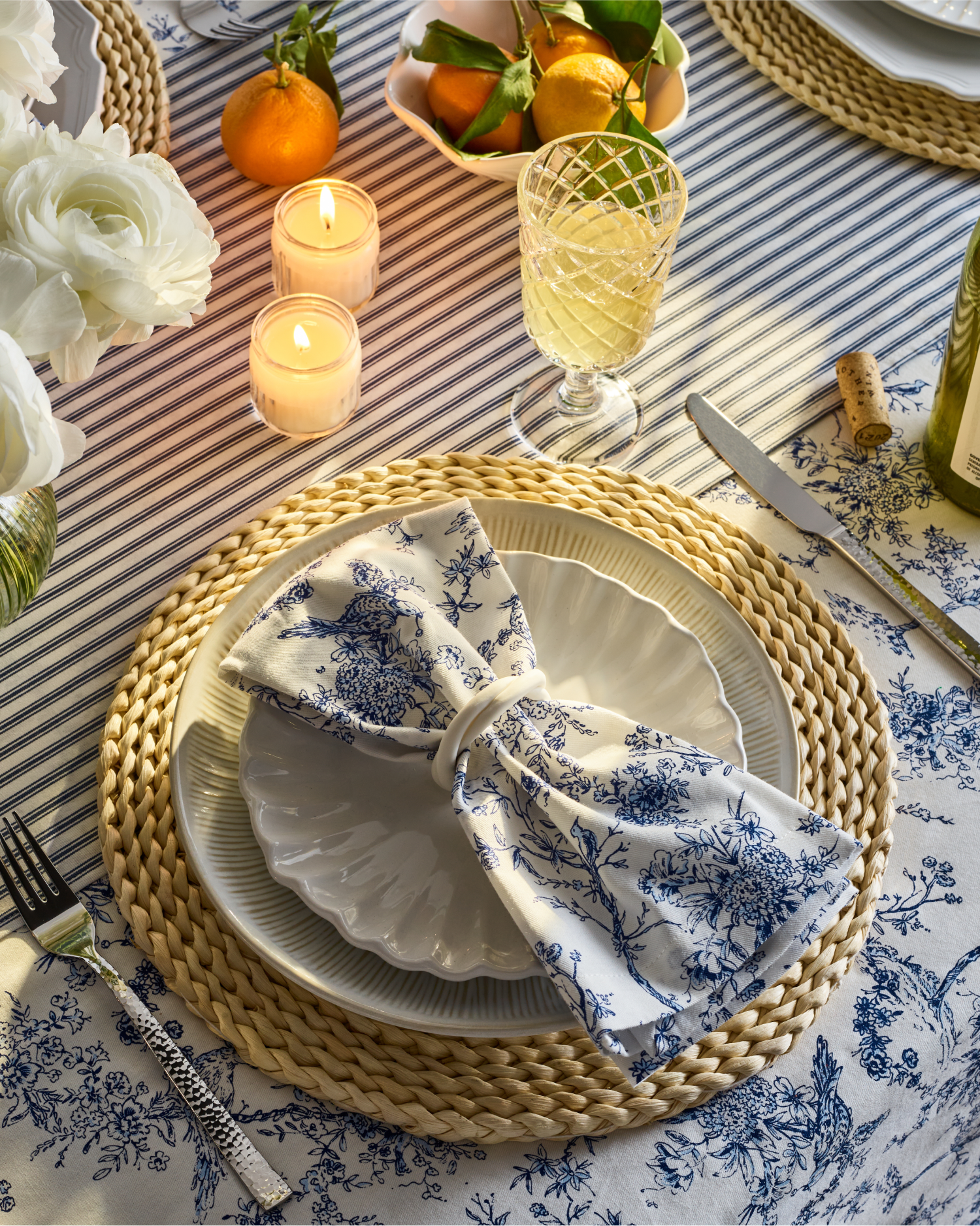 A table setting with a woven placemat, white plates, a floral napkin, wine glass, lit candles, white flowers, and a bowl of oranges on a striped tablecloth, creating an elegant and cozy atmosphere.