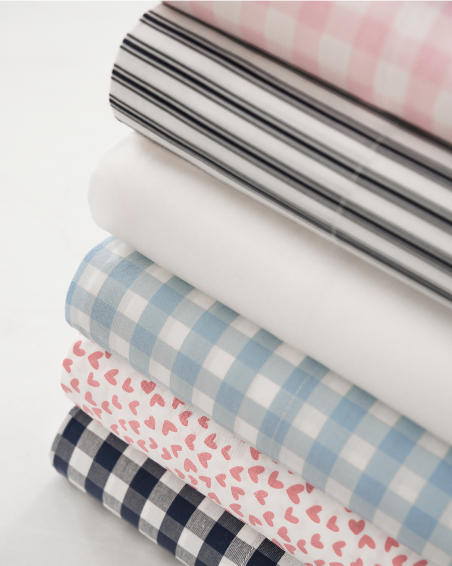 A stack of neatly folded fabrics in various patterns, including black stripes, solid white, blue and white gingham, pink hearts on white, and navy blue and white gingham.