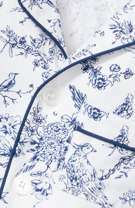 Close-up of a white fabric with blue bird and floral patterns, featuring dark blue piping and white buttons, likely from a shirt or pajama top.