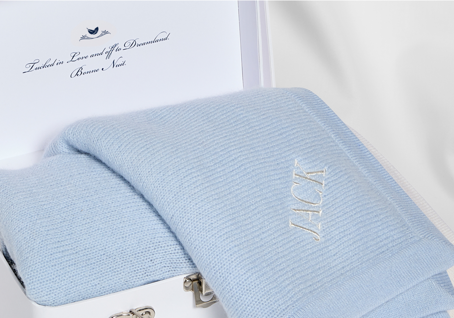 A soft light blue knit blanket embroidered with the name Jack rests inside a white box. The box lid has a printed message that includes the words Tucked in Love and the Dreamland.