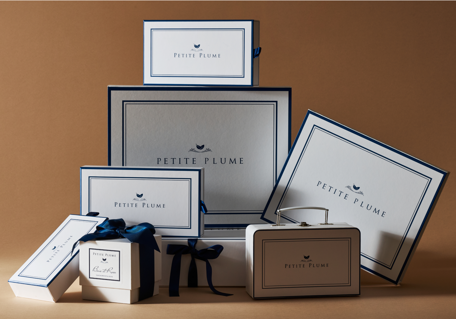 A group of elegant gift boxes in various sizes, all labeled Petite Plume with navy blue borders and ribbon accents, arranged against a neutral brown background.