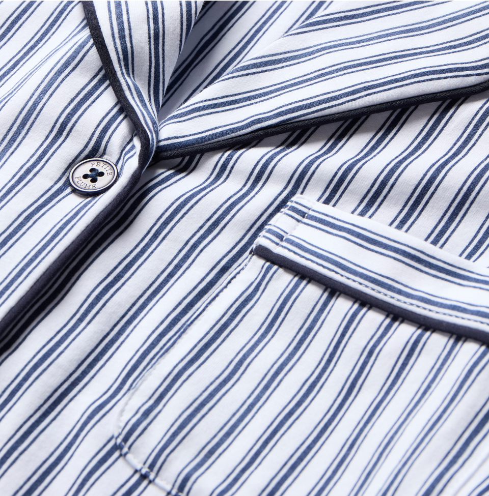 Close-up of a white fabric with navy blue vertical stripes, showing a button, part of a lapel, and a breast pocket, suggesting it is a striped shirt or pajama top.