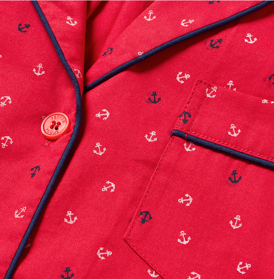 Close-up of a red fabric with small white and navy blue anchor patterns, navy blue piping along the edges, a front pocket, and a red button. The fabric appears to be part of a pajama top or shirt.