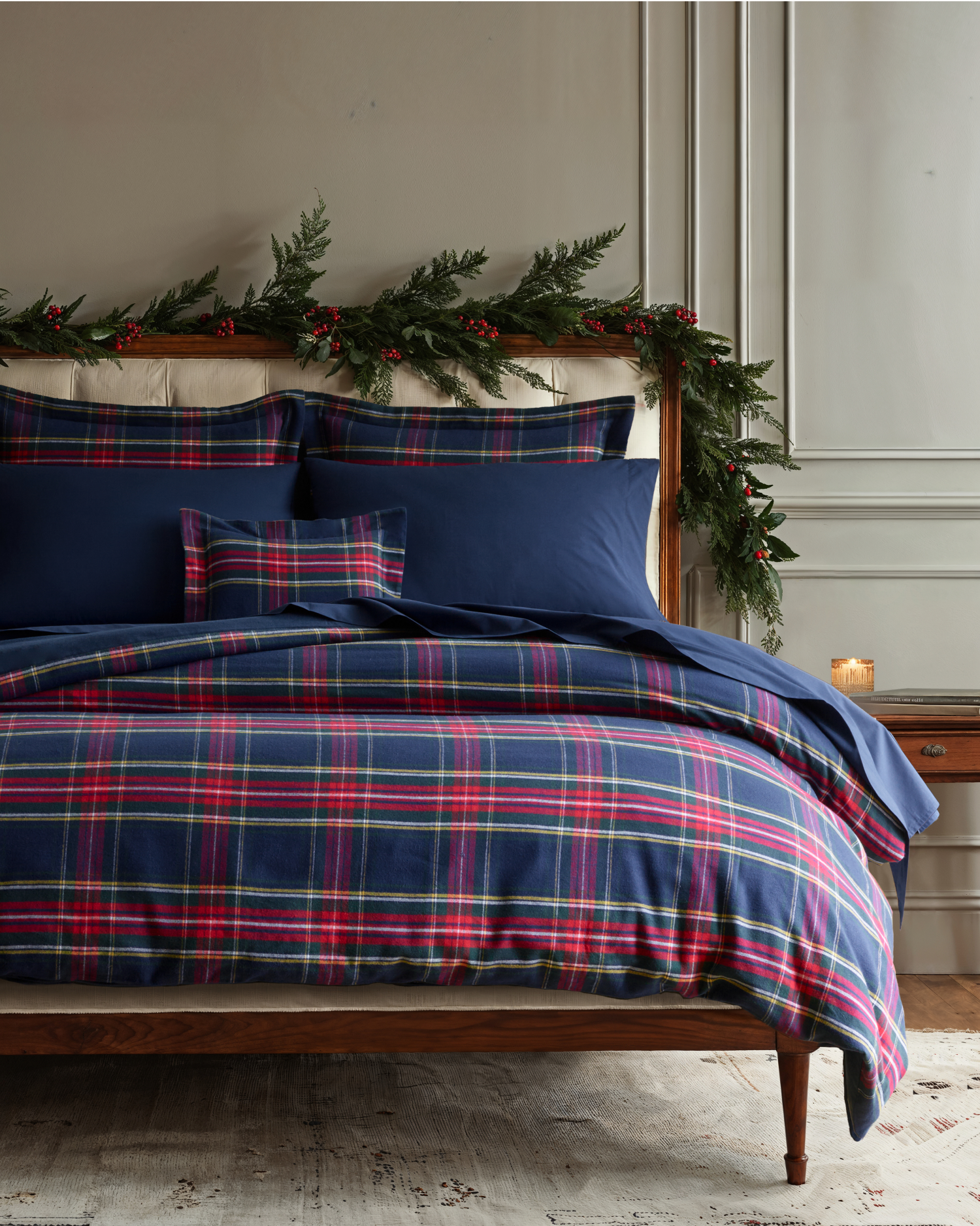 A cozy bed with blue and red plaid bedding, solid navy pillows, and a matching plaid accent pillow. The wooden headboard is decorated with green garland and red berries. A lit candle sits on a bedside table.