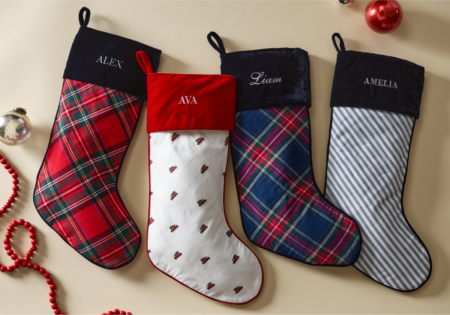 Four personalized Christmas stockings with names Alex, Ava, Liam, and Amelia. The stockings have different festive patterns in red, blue, green, and white, placed on a beige surface with ornaments and red beads nearby.