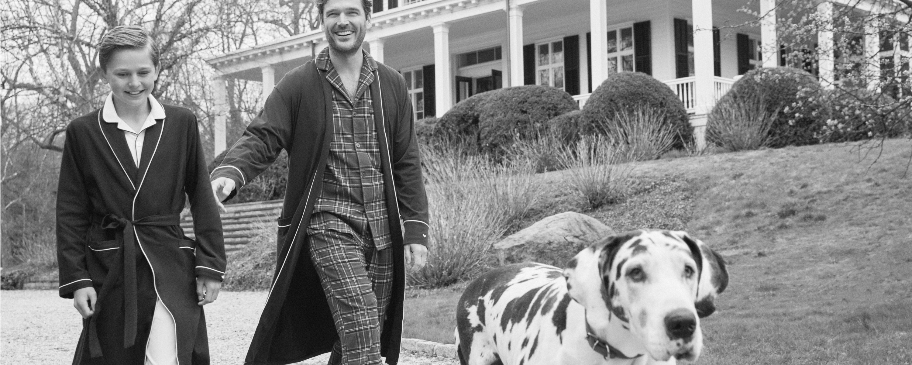 A black-and-white photo of a smiling adult and child, both wearing pajamas and robes, walking outside a large house with a Dalmatian dog in front of them. Bushes and trees are visible in the background. A black-and-white photo of a smiling adult and child, both wearing pajamas and robes, walking outside a large house with a Dalmatian dog in front of them. Bushes and trees are visible in the background.