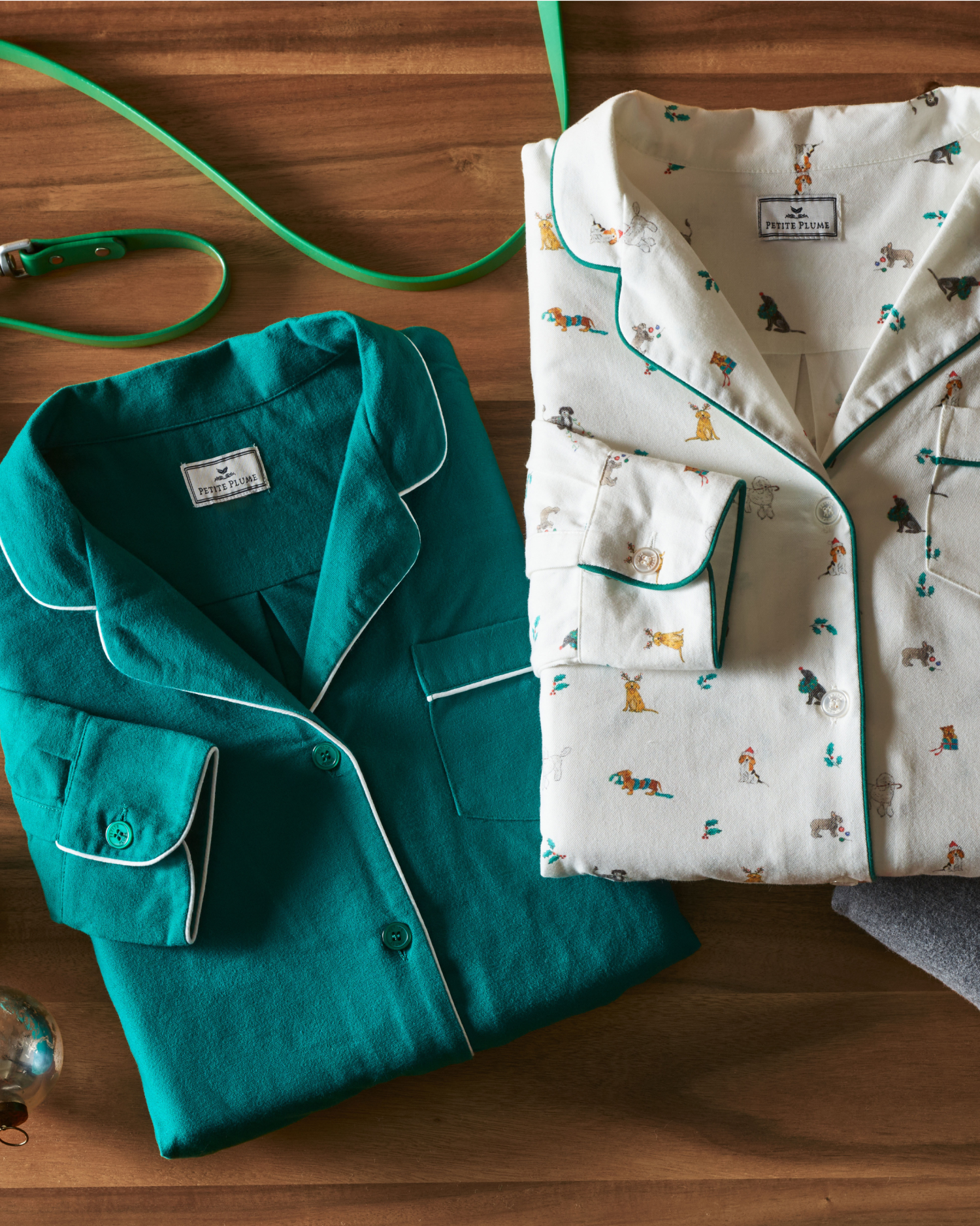 Two folded pajama tops on a wooden surface: one is solid teal with white piping, the other is white with a playful dog print and green trim. Other items, like a green leash and glasses, are partially visible nearby.