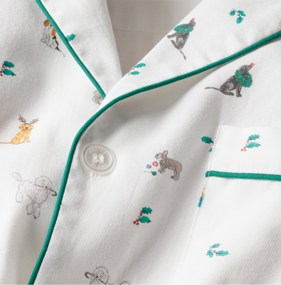 Close-up of a white pajama shirt with green piping, decorated with small illustrations of dogs wearing holiday accessories and scattered holly sprigs. A white button is visible near the center.