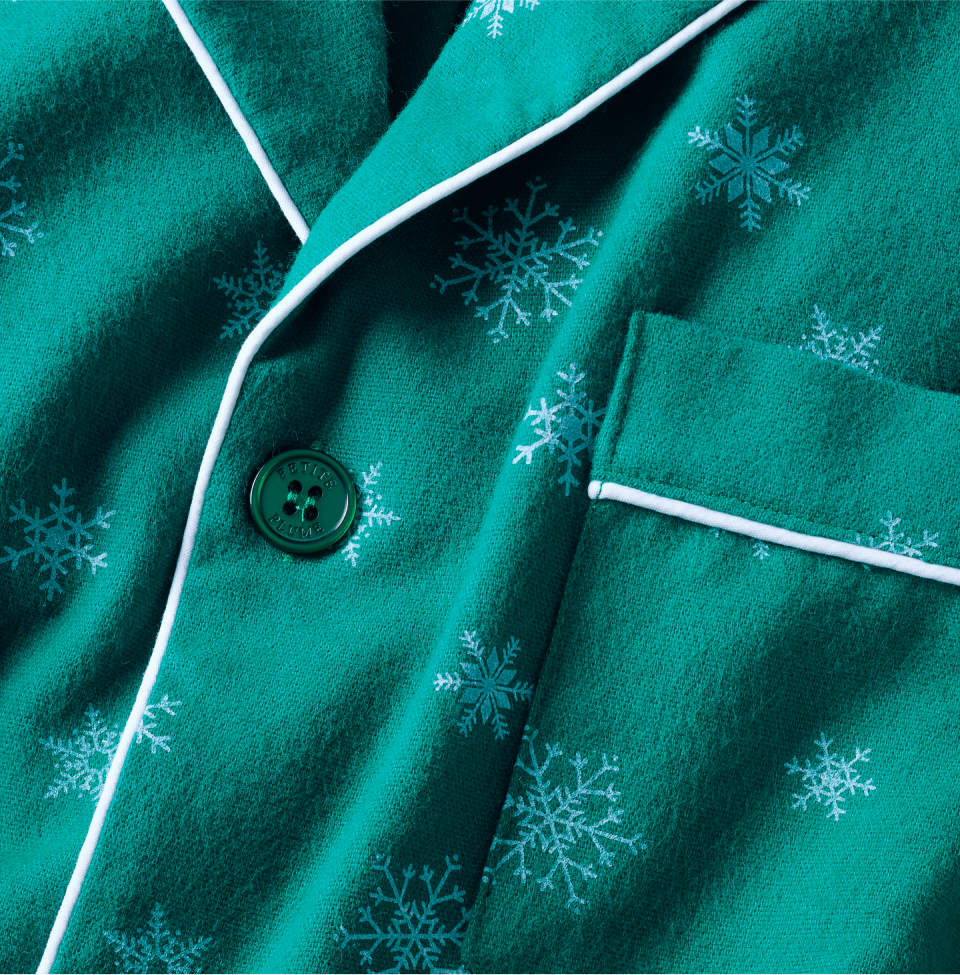 Close-up of green fabric with white snowflake patterns, white piping along the edges, a green button, and a pocket, suggesting it is part of a winter-themed pajama top.