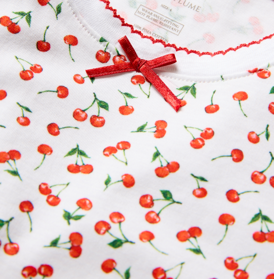 A close-up of a white fabric with a red cherry print, featuring a small red satin bow and red stitching along the neckline.