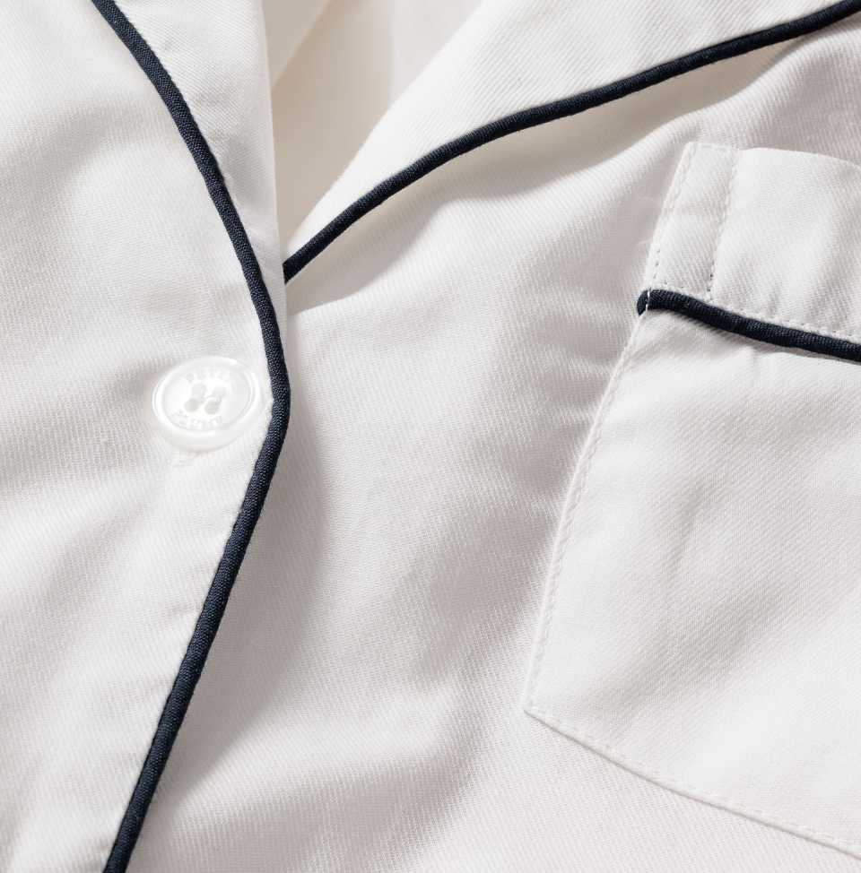 Close-up of a white pajama top with a button, black piping along the collar and pocket, and a visible chest pocket. The fabric appears soft and smooth.
