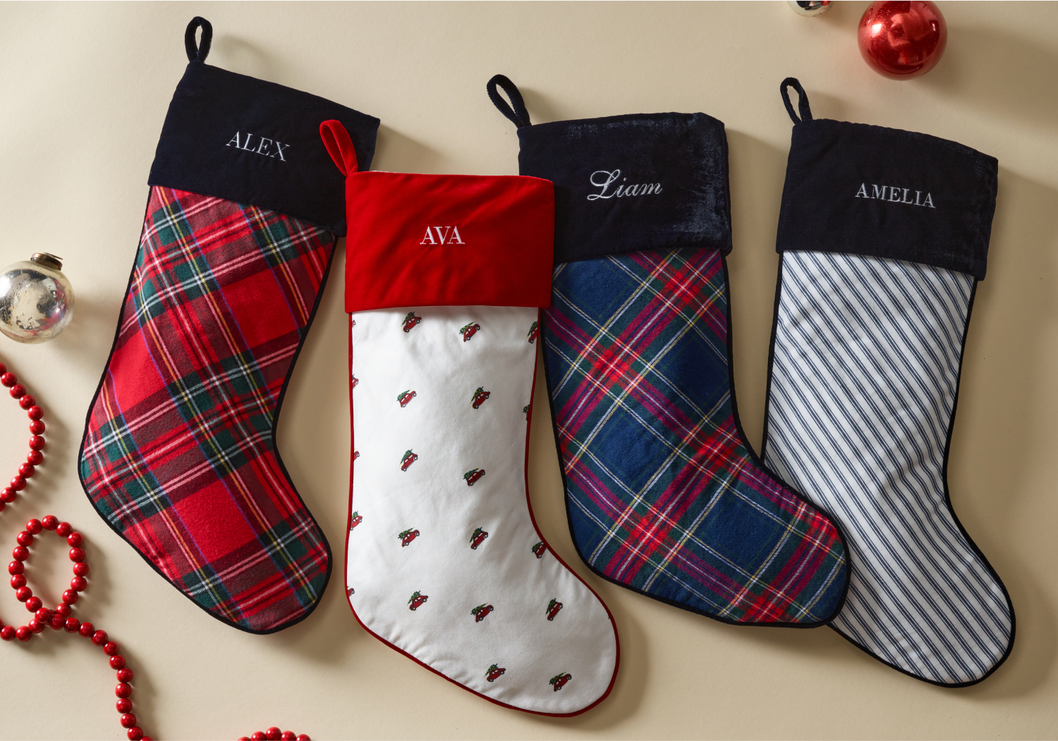 Four Christmas stockings with different patterns and embroidered names—Alex, Ava, Liam, and Amelia—are laid out on a beige surface with red beads and ornaments nearby.