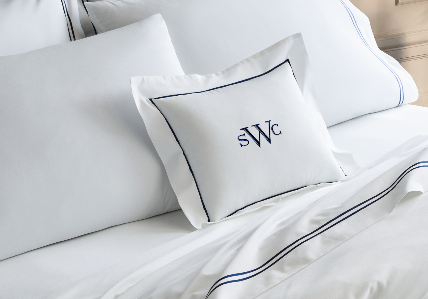 A neatly made bed with crisp white linens and a square pillow featuring dark blue piping and the embroidered initials SWC. The bedding also has subtle blue trim details.