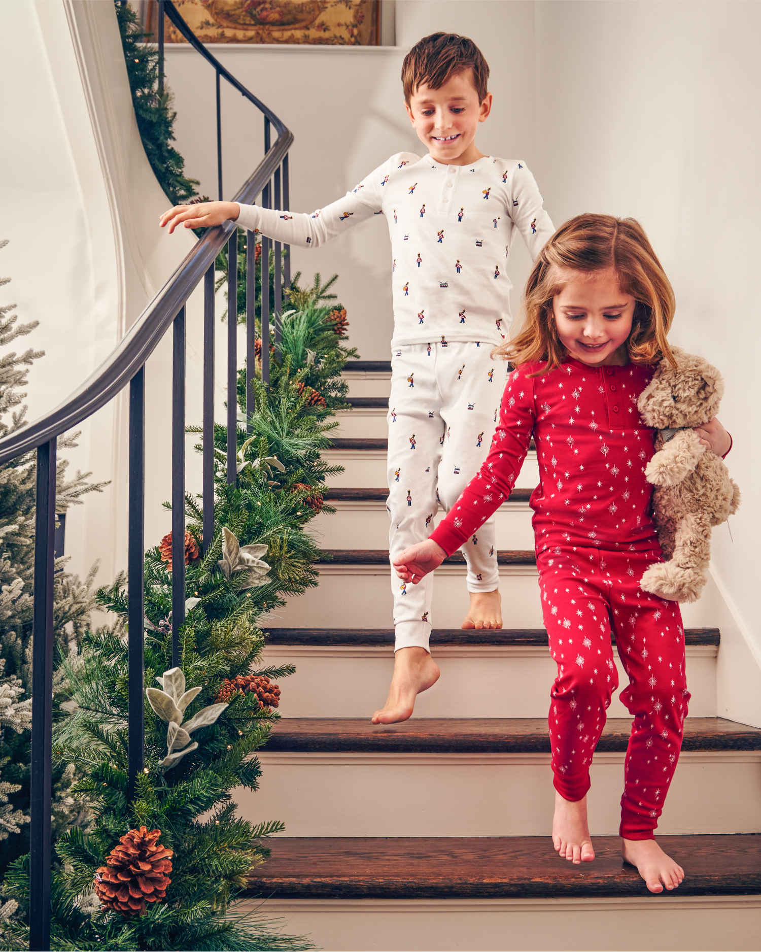 Two children in festive pajamas, one in white and one in red, walk down a decorated staircase with pine garlands. The girl holds a teddy bear and both are smiling, suggesting a joyful holiday morning.