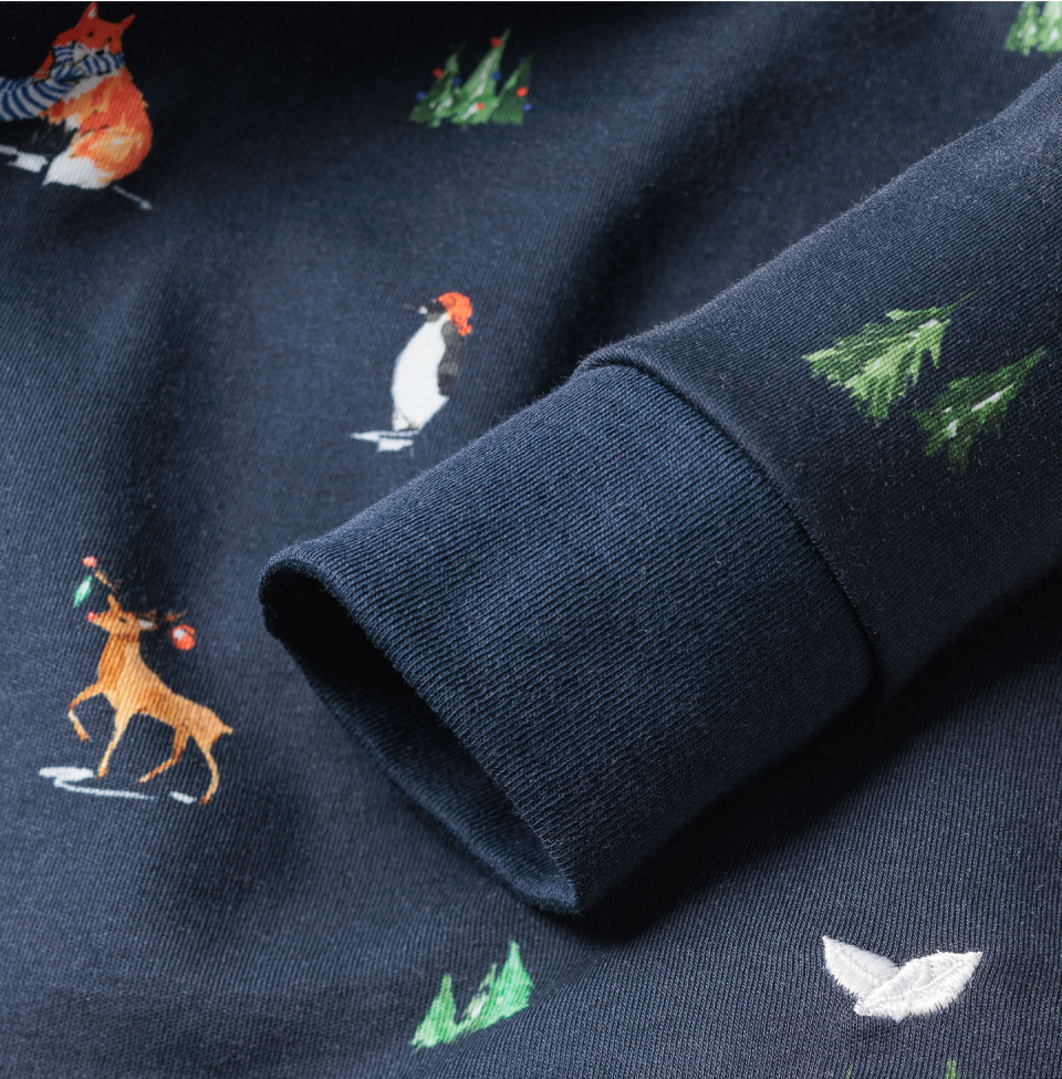 A close-up of a navy blue fabric with embroidered illustrations of a penguin, a scarf-wearing fox, a reindeer with lights, green pine trees, and a white bird, next to a folded sleeve.