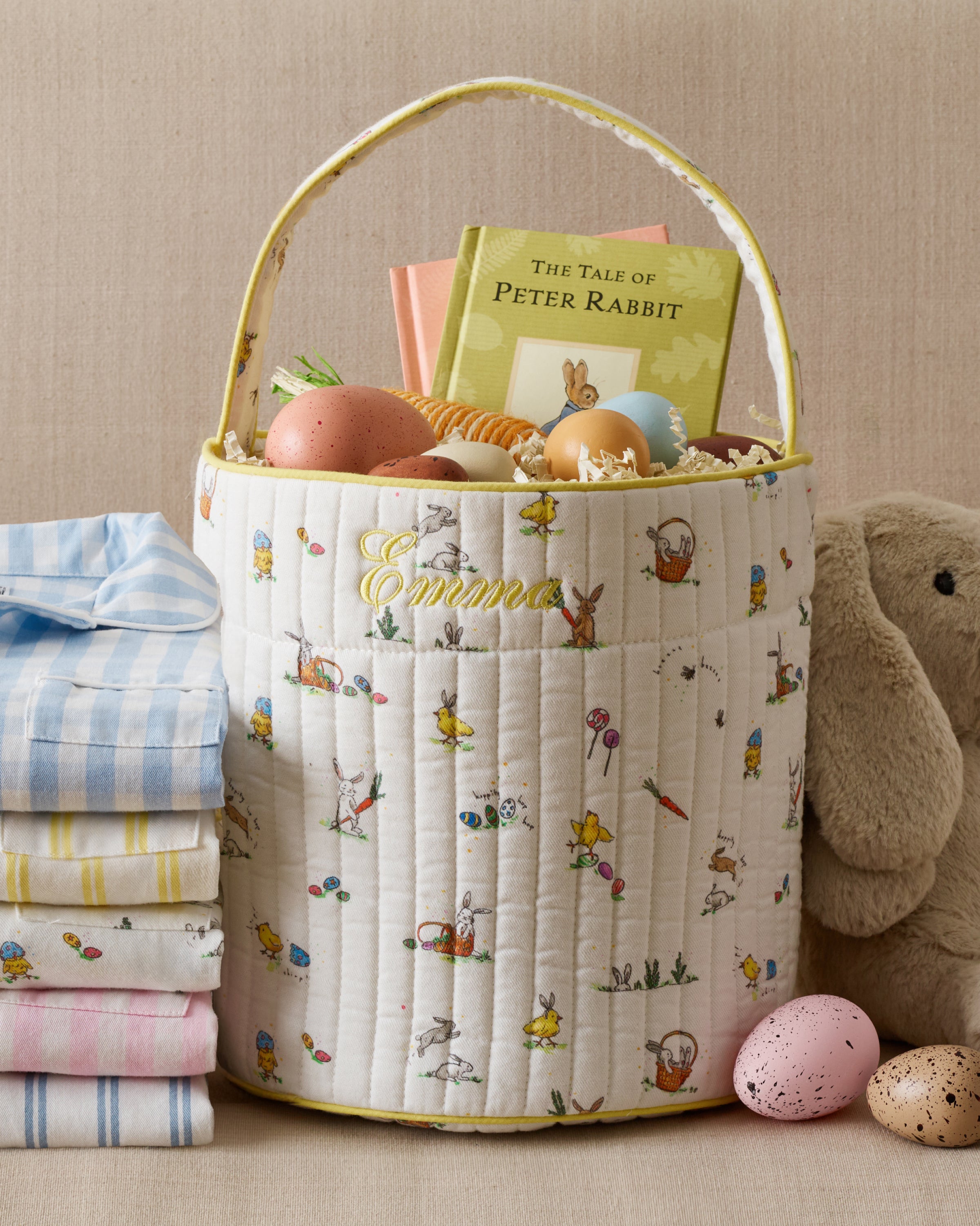 A Kid's Quilted Easter Basket personalized with "Emma," filled with decorated eggs, a Peter Rabbit book, and a toy, sits next to pastel linens, a plush bunny, and speckled eggs on a beige surface.