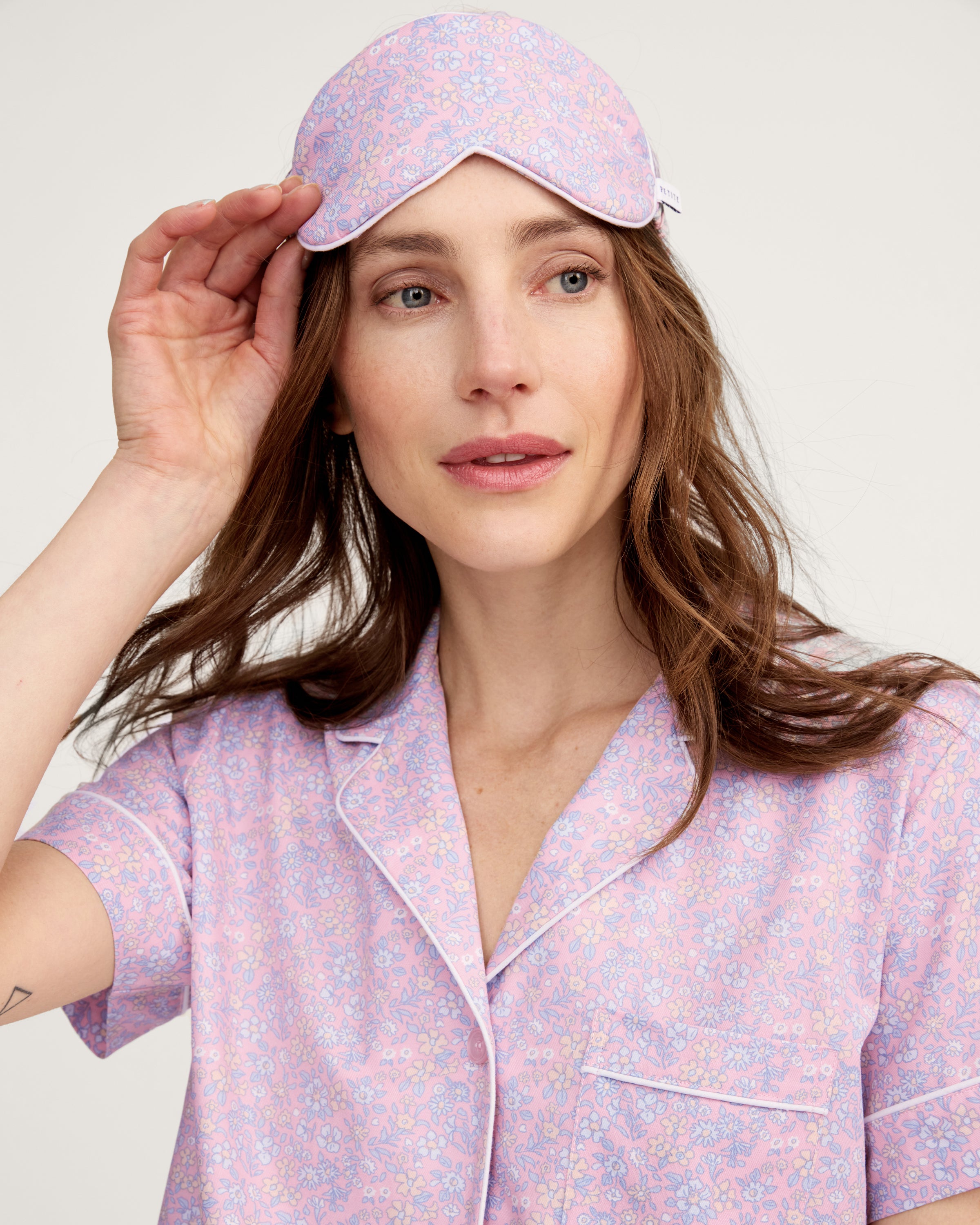 A woman with long brown hair wears the Adult's Twill Sleep Mask in Fleur Blush, lifting it slightly with one hand as she gazes off to the side with a relaxed expression.