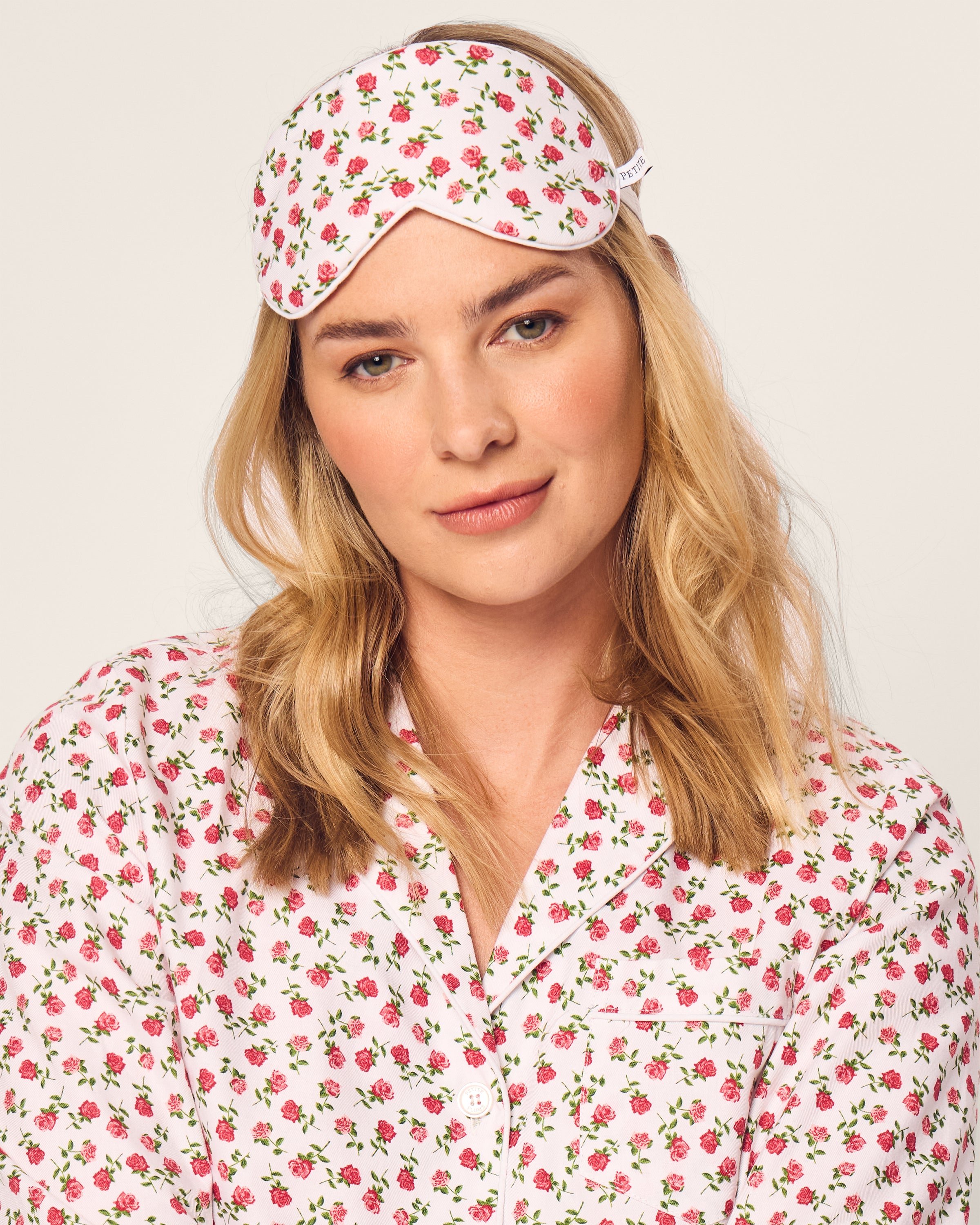 A woman stands against a plain backdrop, wearing a monogrammed floral pajama set with pink and green roses on white. Her light-colored hair complements the matching Adults Twill Sleep Mask in Petite Petals by Petite Plume, adding elegance to her cozy ensemble.