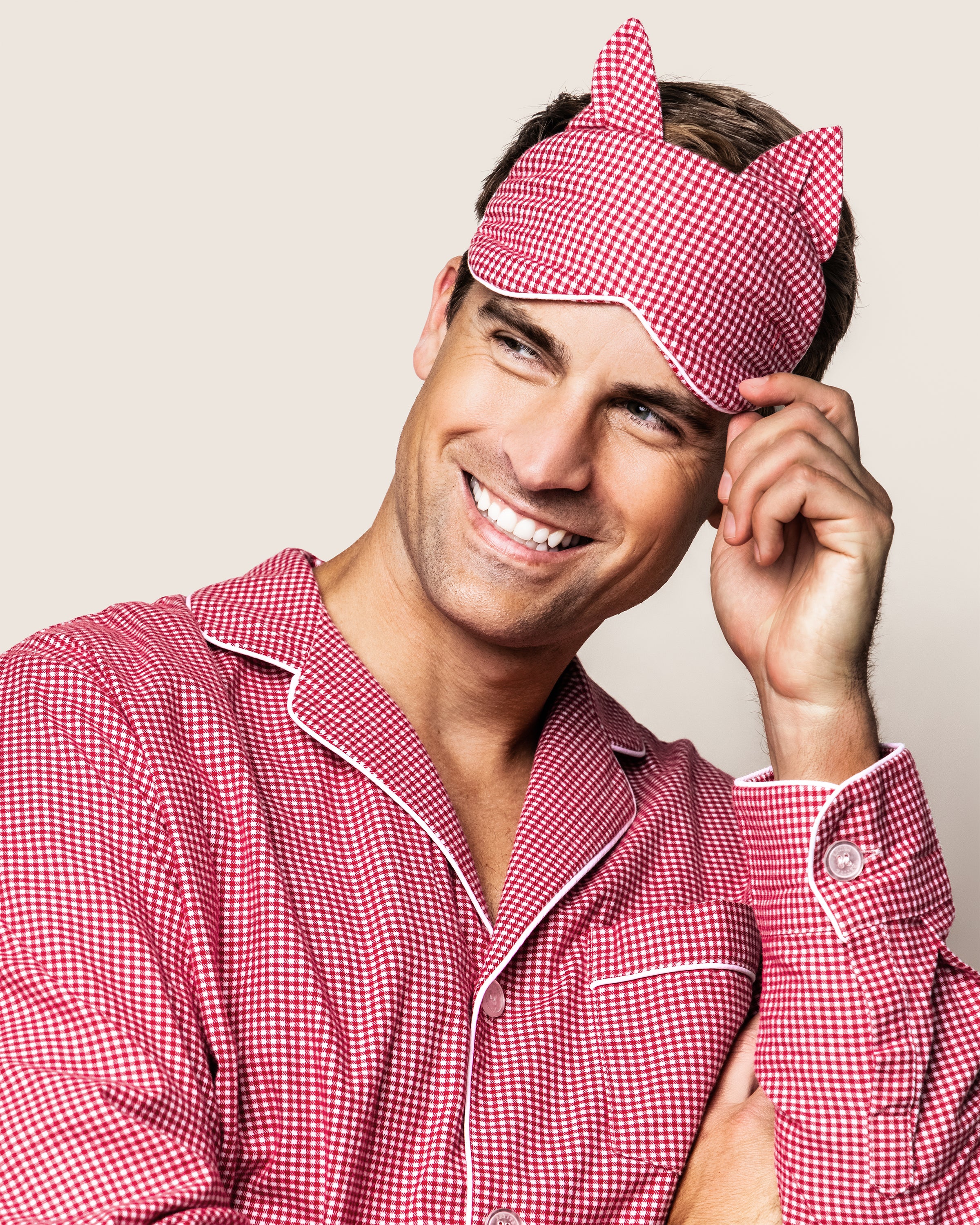 A man smiles as he adjusts the Petite Plume Adults Flannel Kitty Sleep Mask in Red Mini-Gingham, paired with matching luxury sleepwear. Hes gazing to the side against a plain, light background.