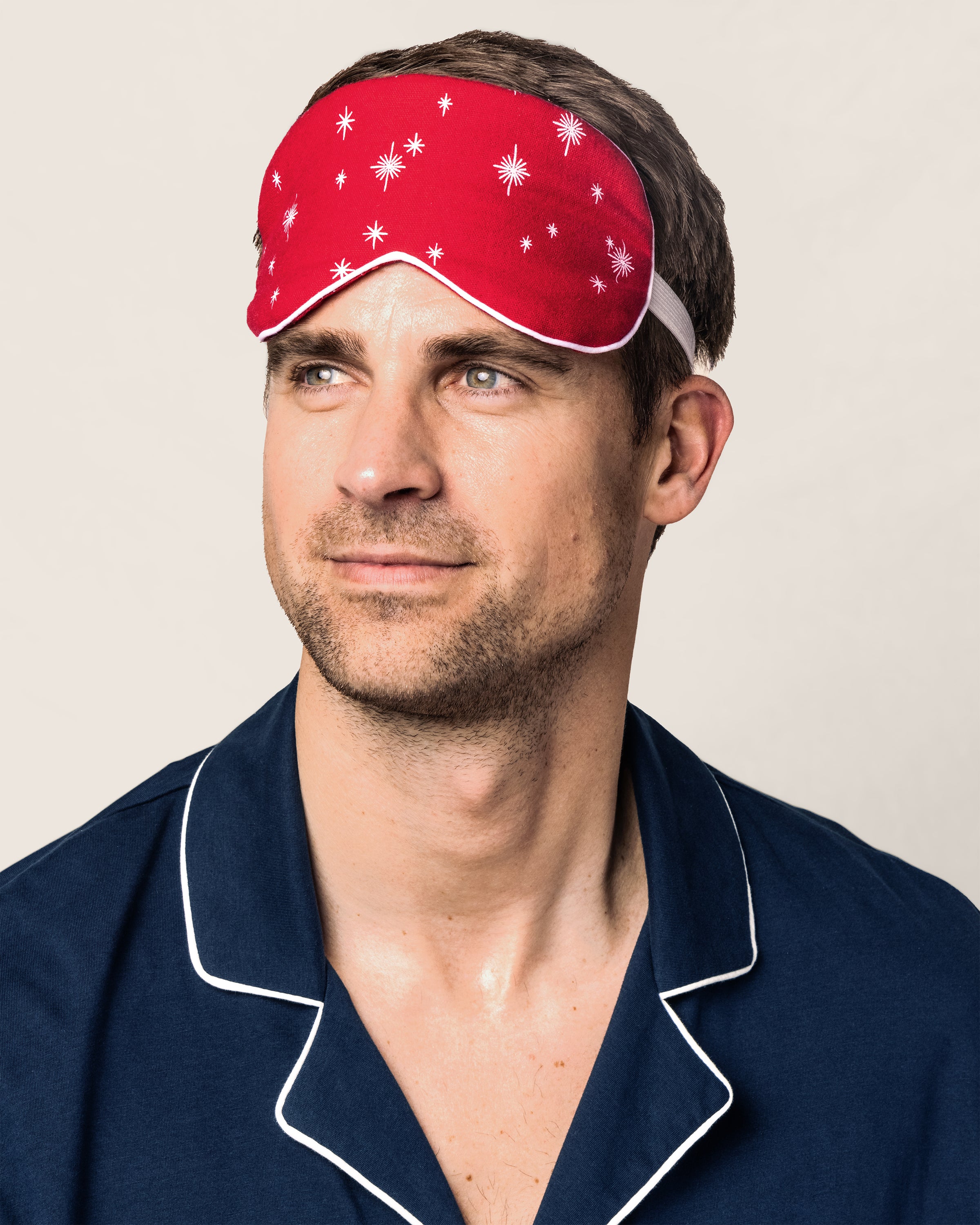 A man in navy pajamas with white trim and a red sleep mask featuring white stars looks aside against a simple backdrop, displaying Petite Plume’s Adults Flannel Sleep Mask in Starry Night, a perfect gift for any occasion.