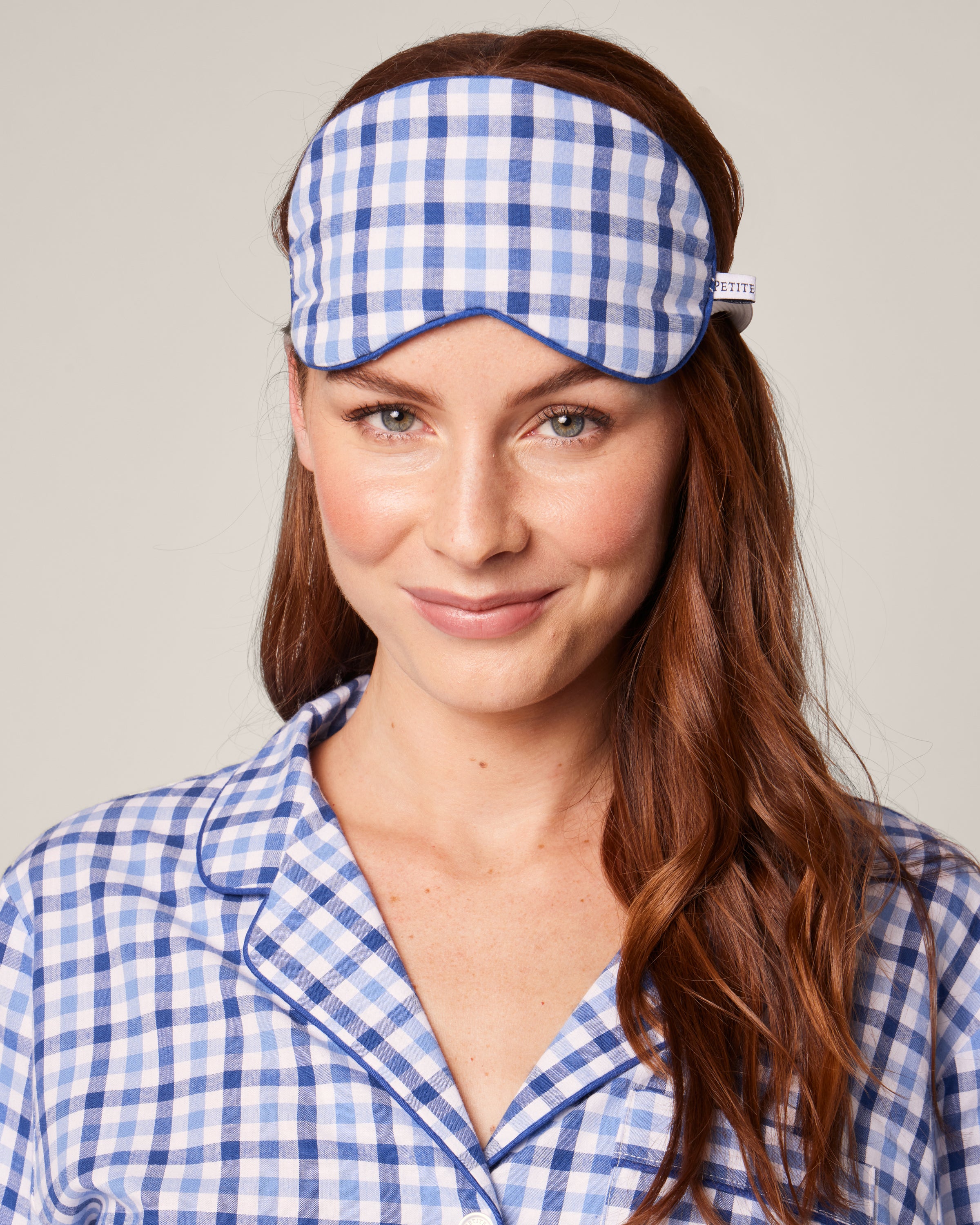 A woman with long auburn hair wears a blue and white checkered pajama set with Petite Plumes Adults Twill Sleep Mask in Royal Blue Gingham, featuring monogramming, on her head. She smiles warmly at the camera against a plain background.