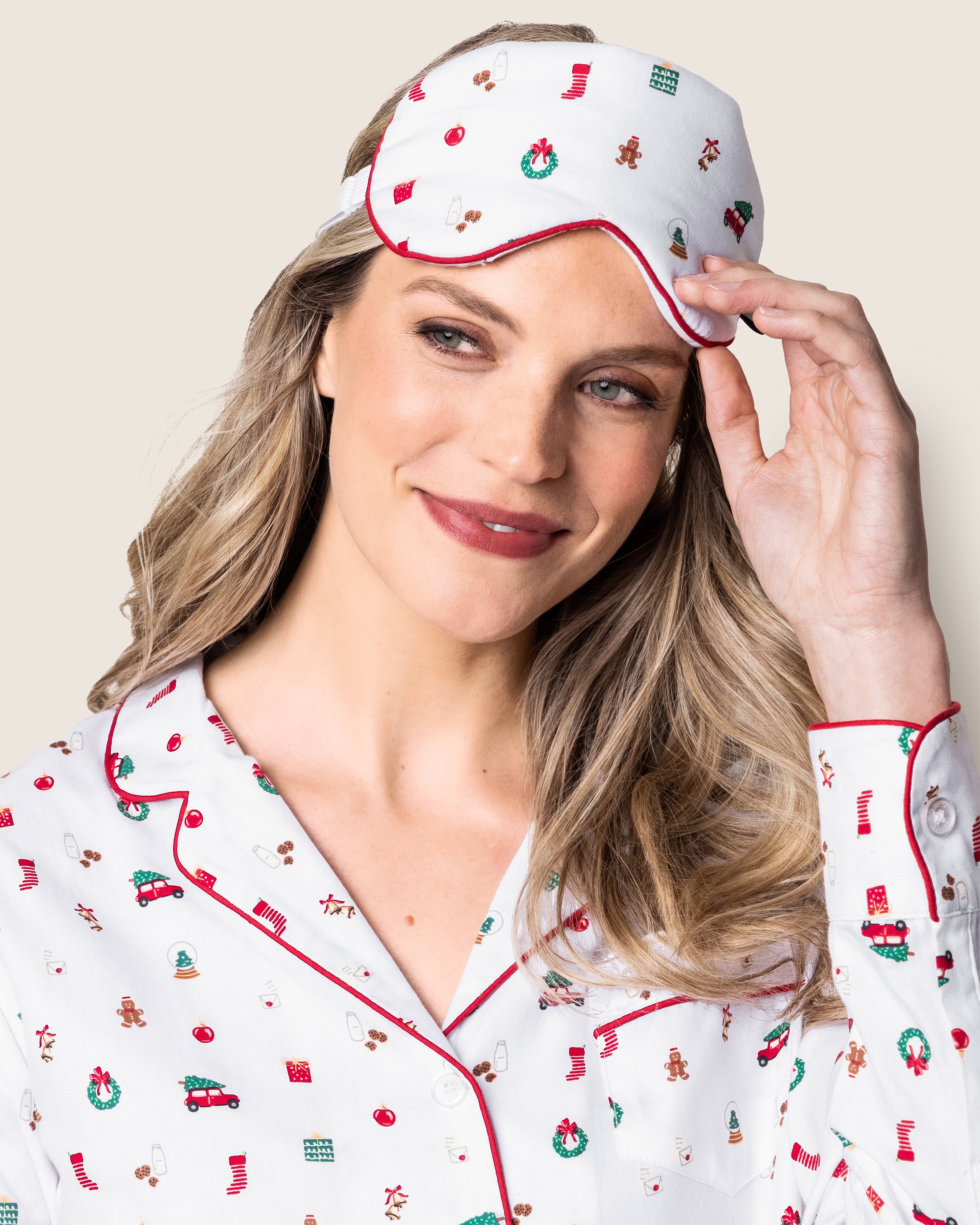 A person beams in festive white pajamas with red piping, featuring gingerbread men and wreaths. They enhance the luxe look with Petite Plumes Adults Twill Sleep Mask in Winter Nostalgia. Their long, wavy hair flows elegantly as they pose against a plain backdrop.