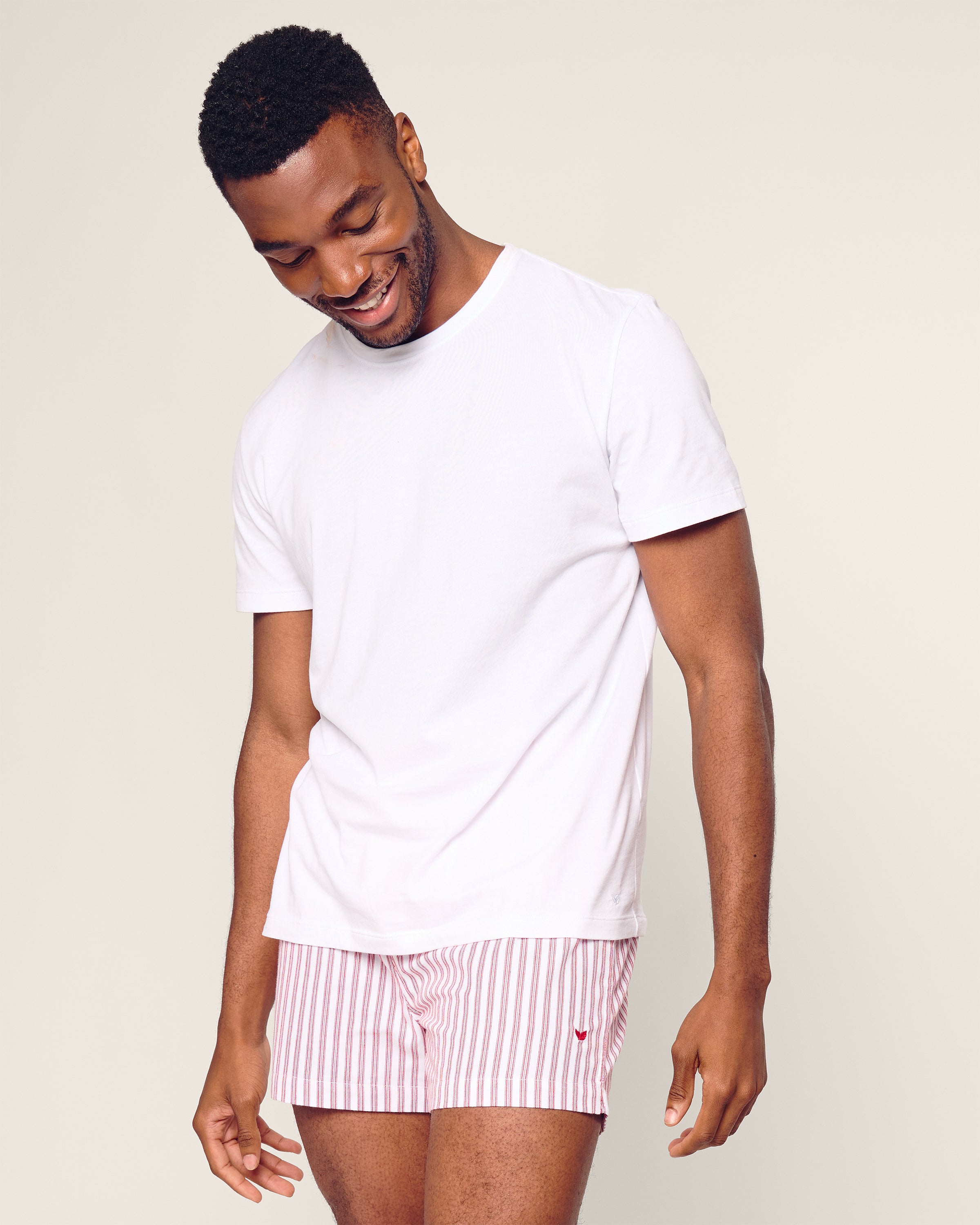 A man in a plain white T-shirt and Petite Plumes Mens Twill 3 Pack Boxers in Holiday Red smiles while looking downward against a light, neutral background.