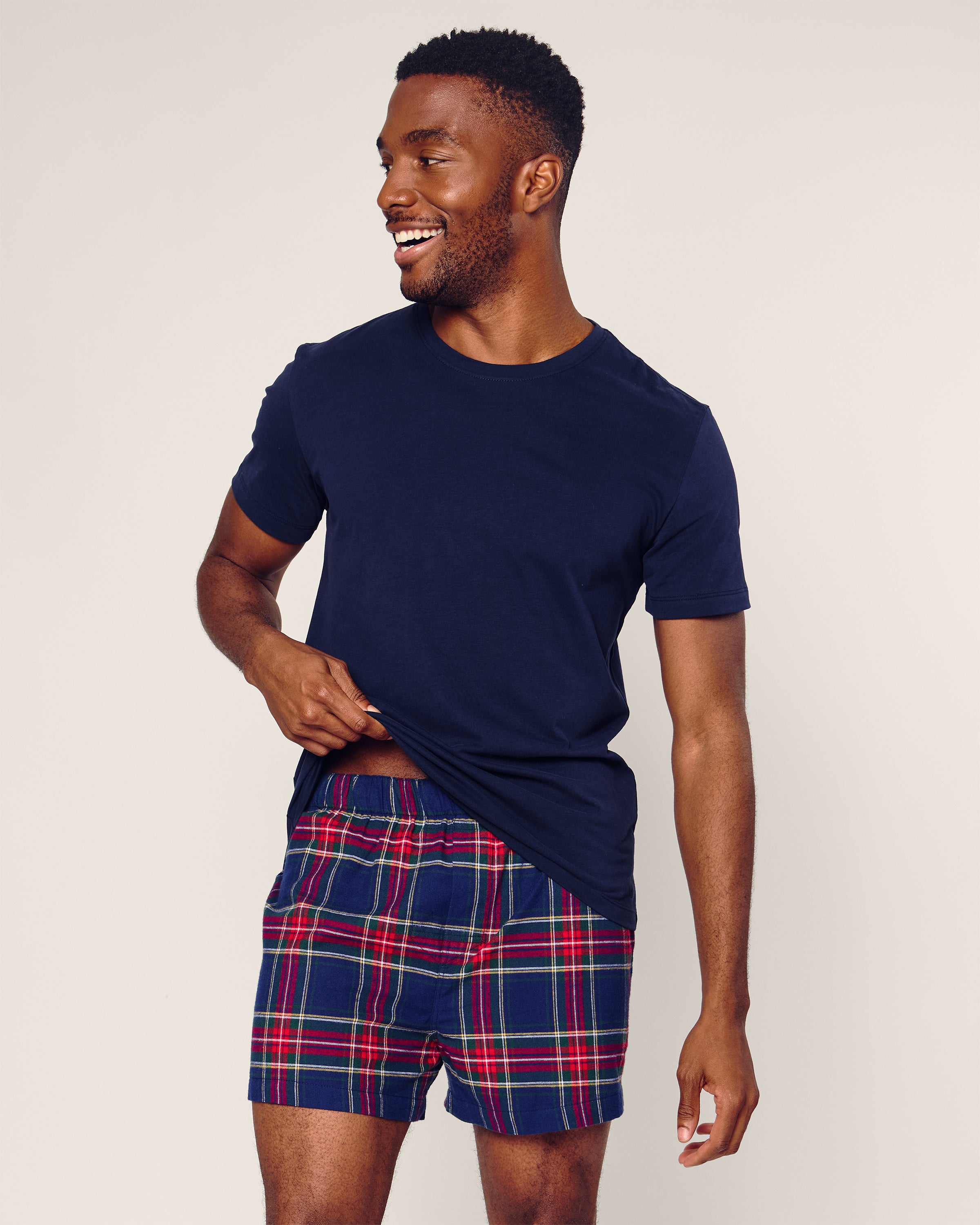 A man in a navy blue t-shirt and Petite Plume Mens Twill 3 Pack Boxers in Windsor Tartan smiles, looking to the side while lifting part of his shirt against a plain light background.