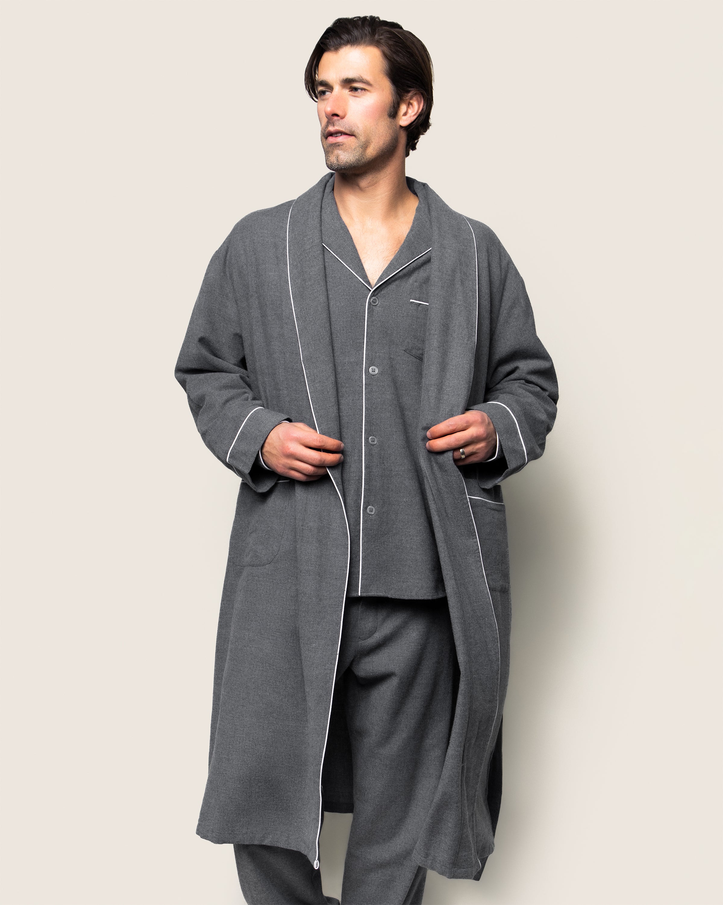 A man stands against a plain backdrop, dressed in Petite Plumes Mens Flannel Robe in Grey with matching pajamas. The robe, made from 100% cotton, showcases elegant white piping. With one hand in his pocket, he gazes to the side.