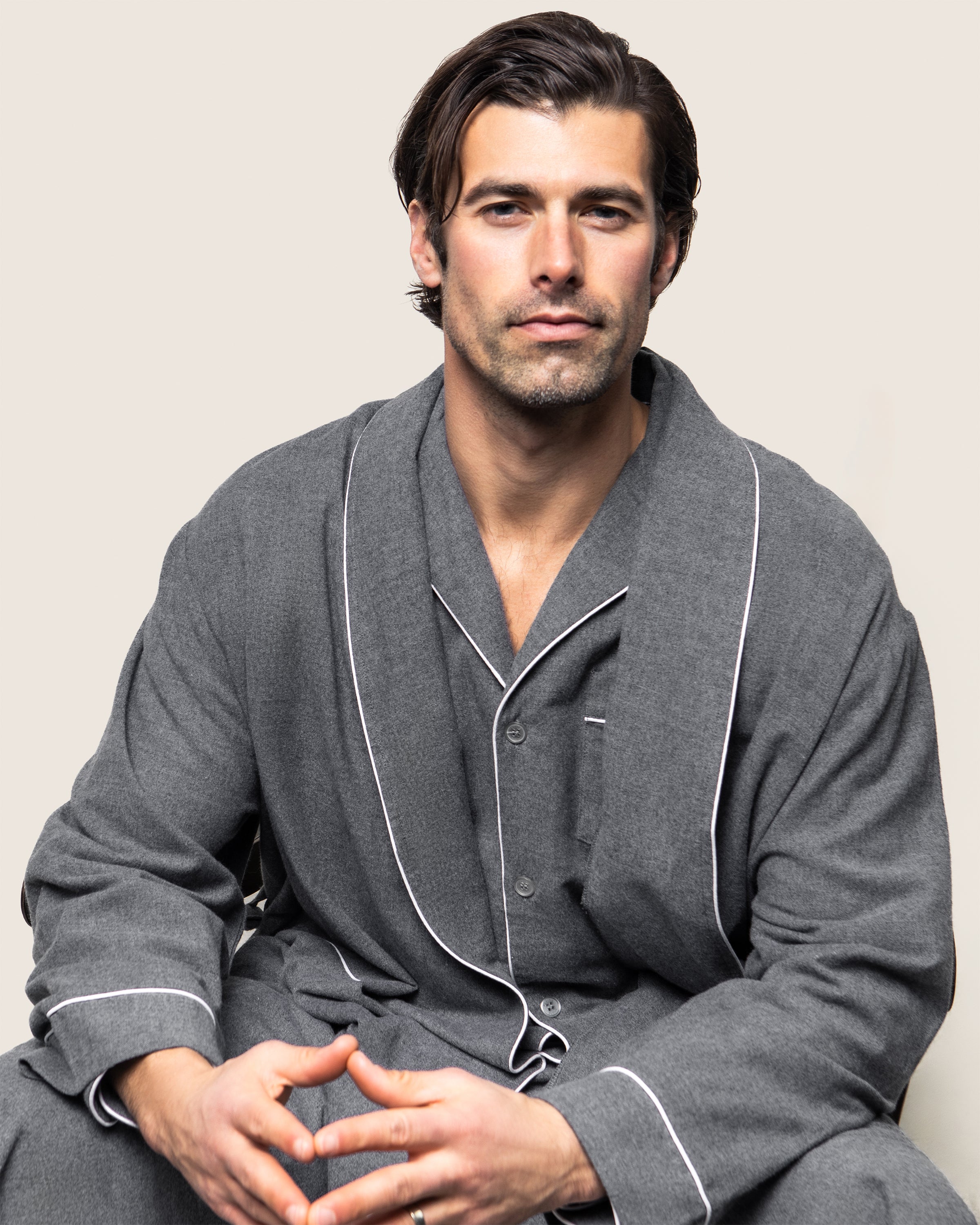A man with short brown hair sits against a plain background, wearing Petite Plumes Mens Flannel Robe in Grey. He has a serious expression and his hands are loosely clasped in front of him.