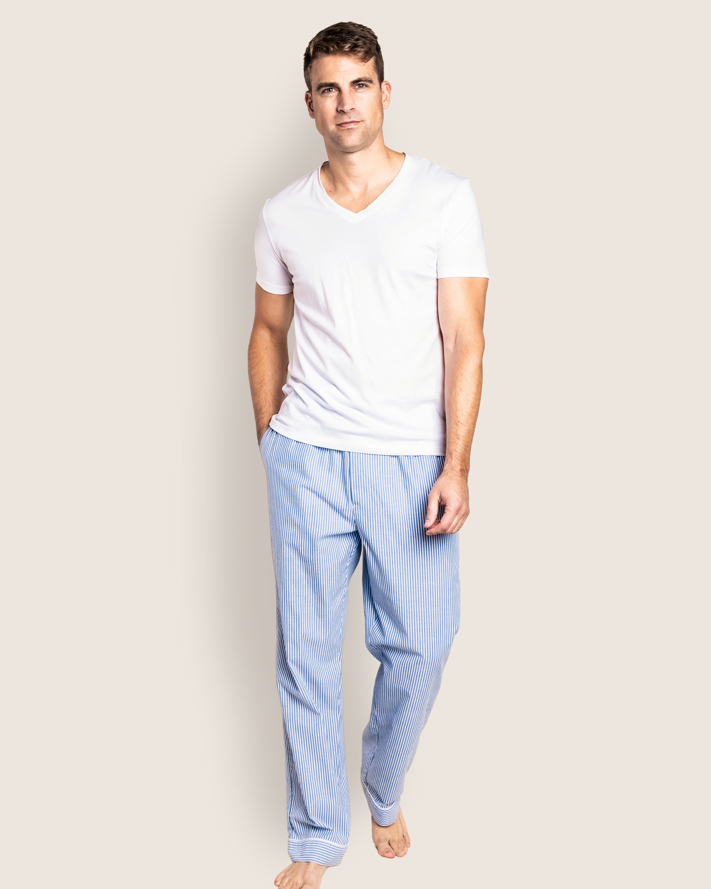 A person stands barefoot against a plain background, hand in pocket, wearing a white V-neck T-shirt and Petite Plumes Mens Twill Pajama Pants in French Blue Seersucker, expertly crafted from the finest quality cotton.
